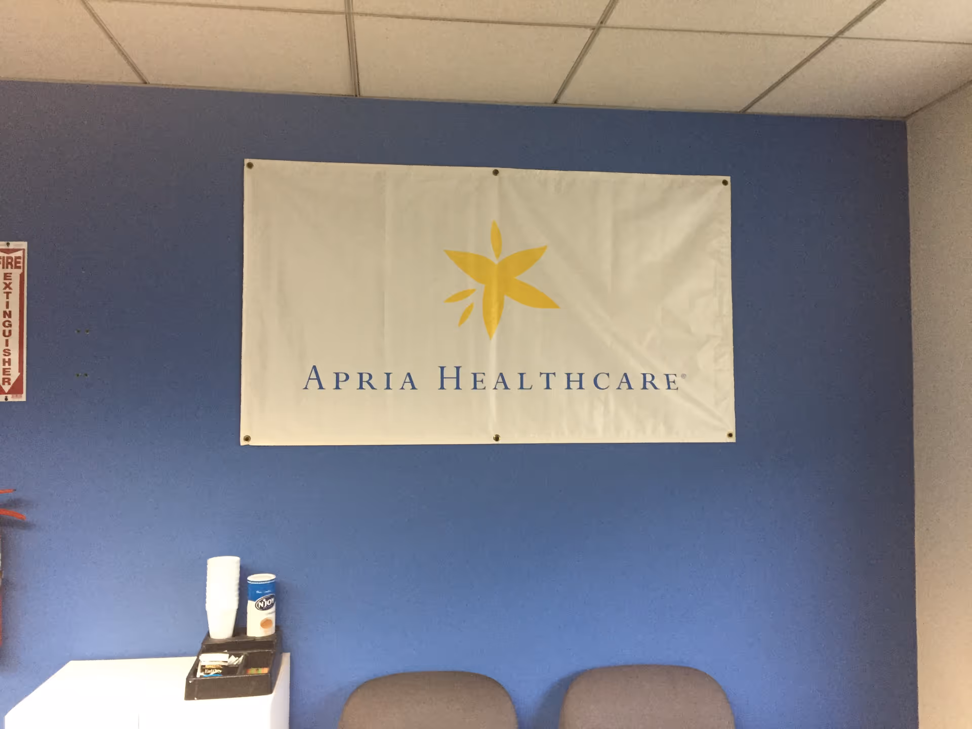 Blue interior wall with Apria Healthcare banner, two chairs, and a small counter.