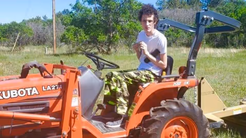 Person sits on orange tractor in a grassy, tree-filled outdoor area.