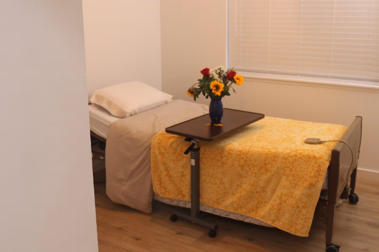A bedroom with a bed, yellow patterned blanket, a rolling tray, and a vase of flowers.