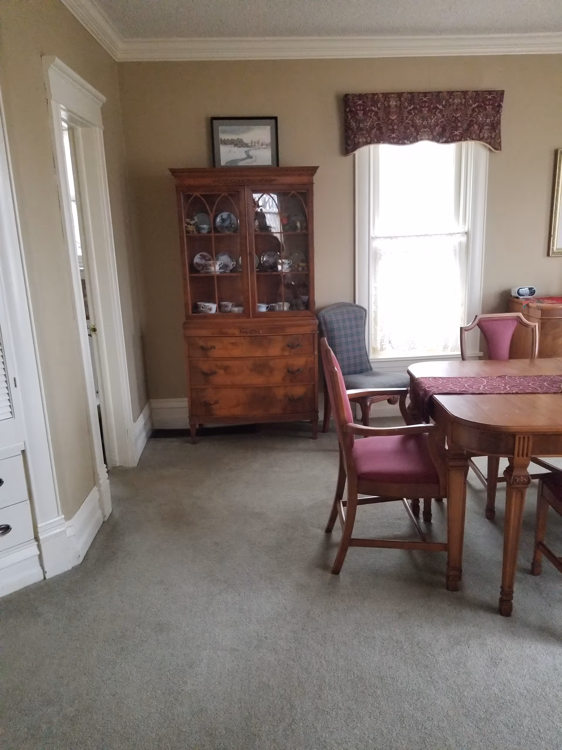 Traditional dining room with a wooden table, chairs, china cabinet, window, and beige carpet.