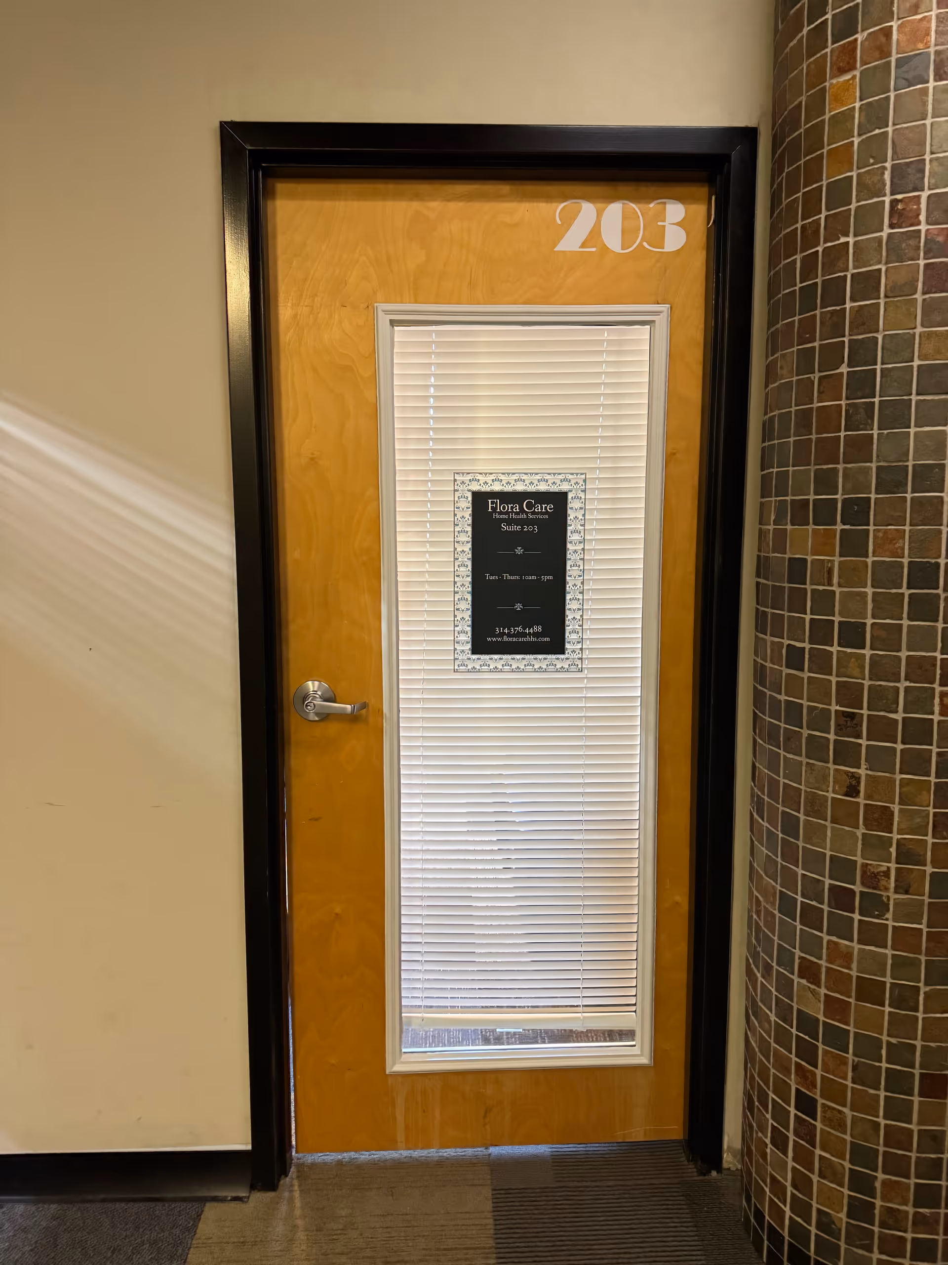 Interior hallway door labeled 203 for Flora Care, with blinds and a curved tiled column.