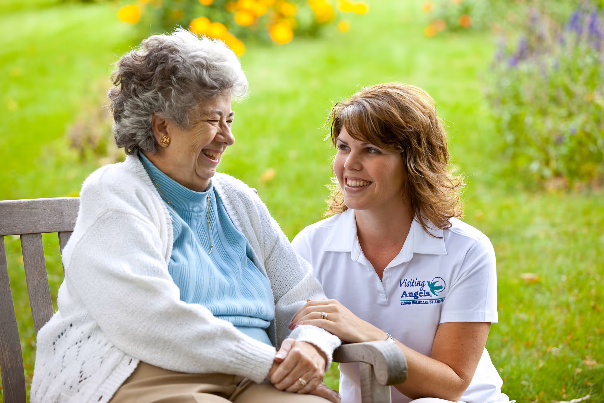 A caregiver and senior smile while seated on a bench in a sunny garden.