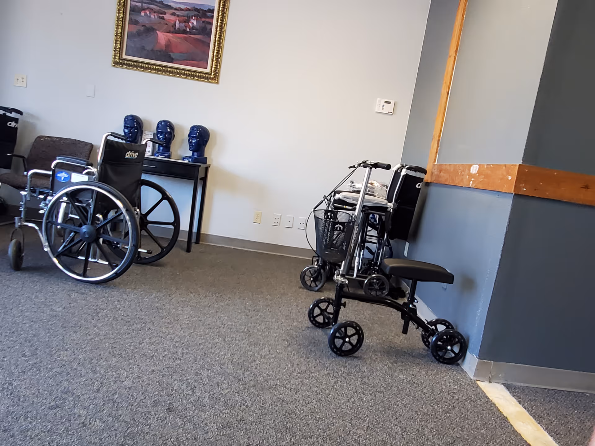 Wheelchair, walker, and medical equipment in a quiet facility hallway, with a chair and painting.