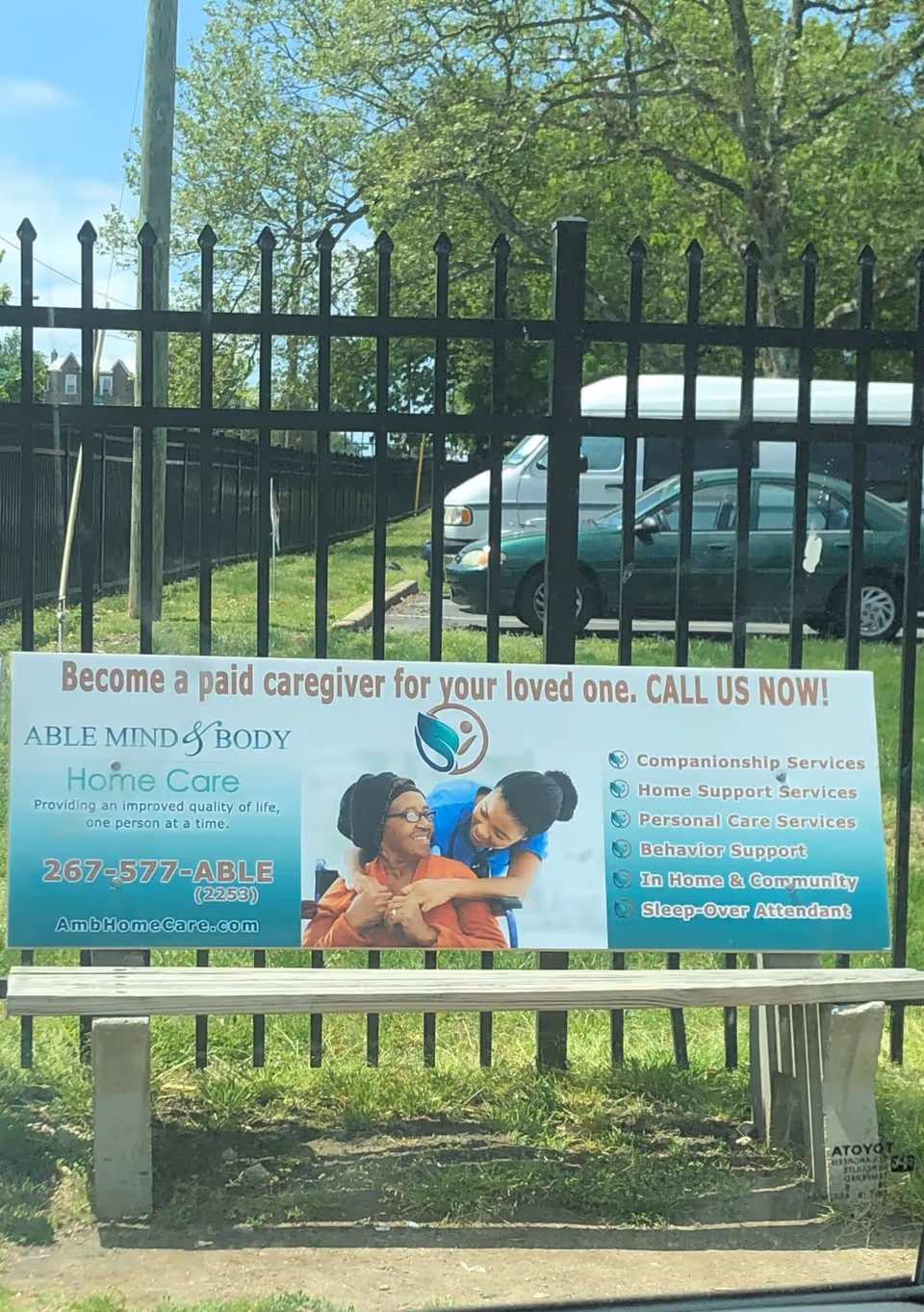 Outdoor fence banner promoting Able Mind & Body Home Care beside a bench.
