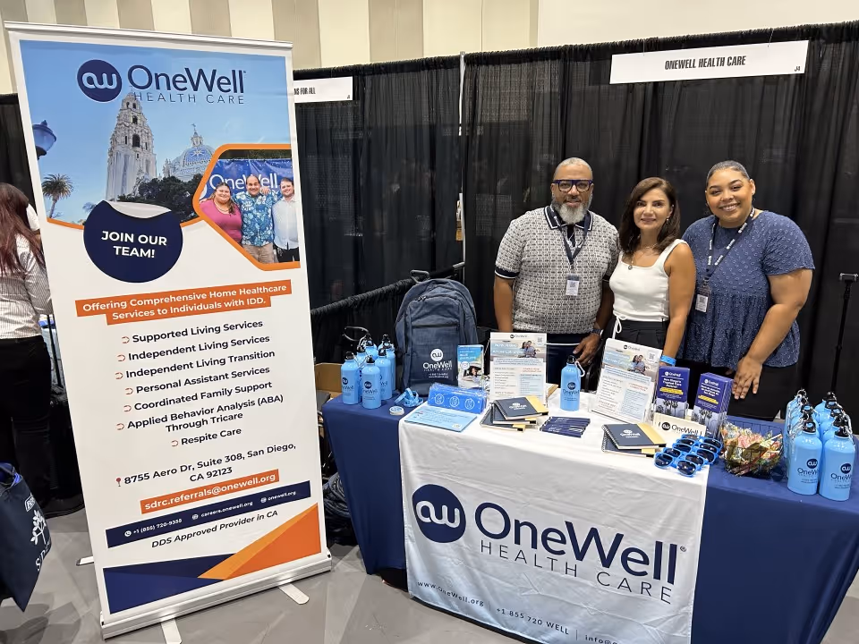 Three staff members stand behind a OneWell Health Care booth at a trade show.