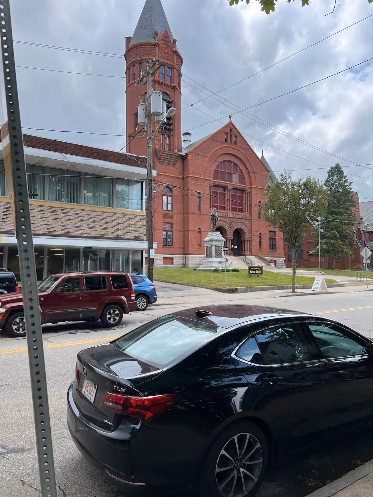 Red brick building with arched windows and a tall tower beside a street with parked cars.