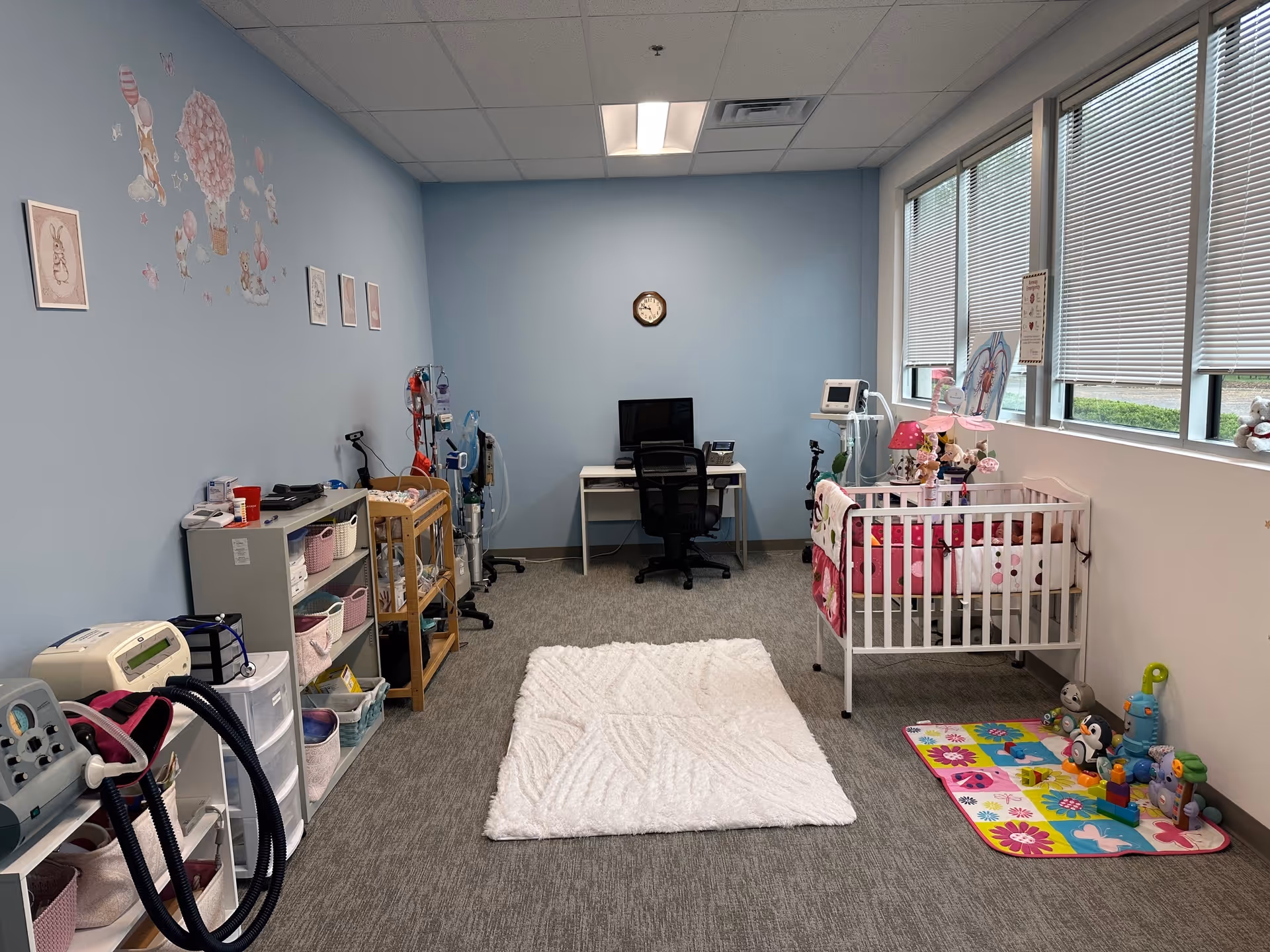 Bright pediatric room with a crib, toys, medical equipment, desk, and blue walls.