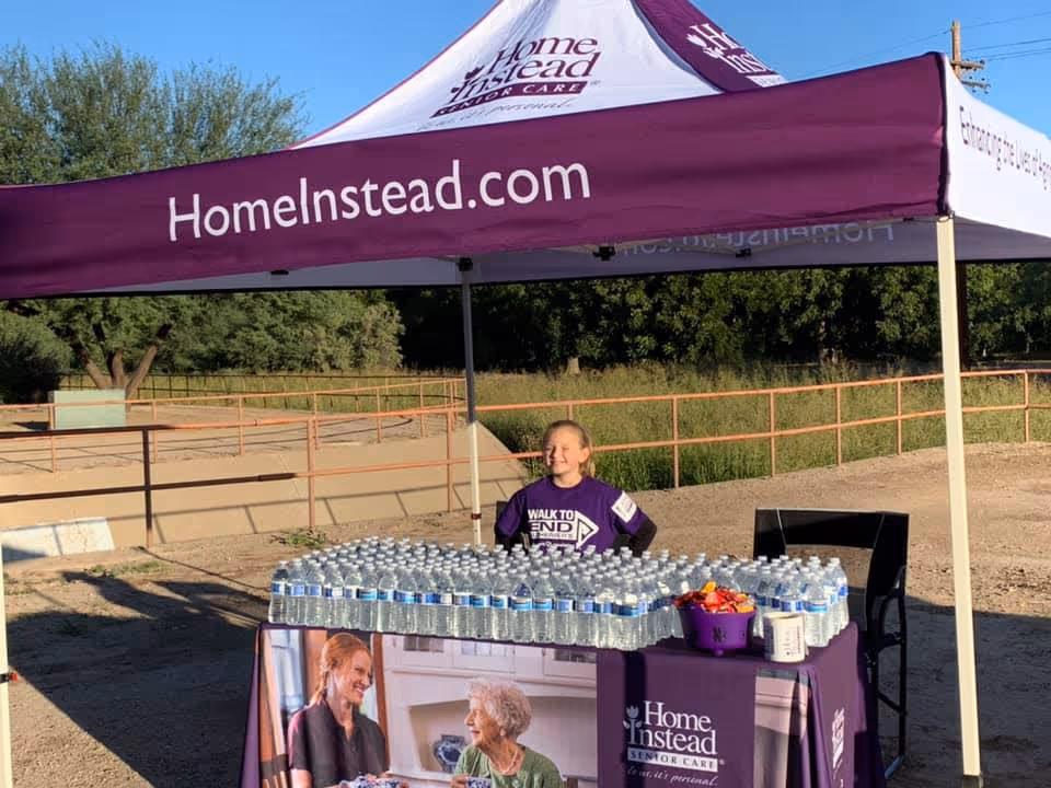 Purple Home Instead tent at an outdoor event with bottled water on a table.