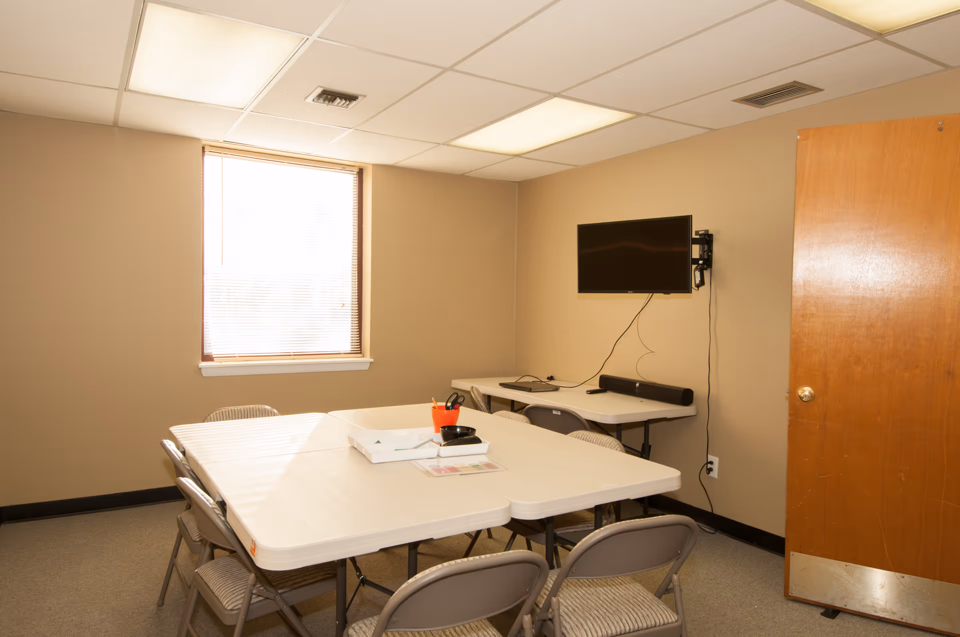 A small beige meeting room with folding tables, chairs, and a wall-mounted TV.