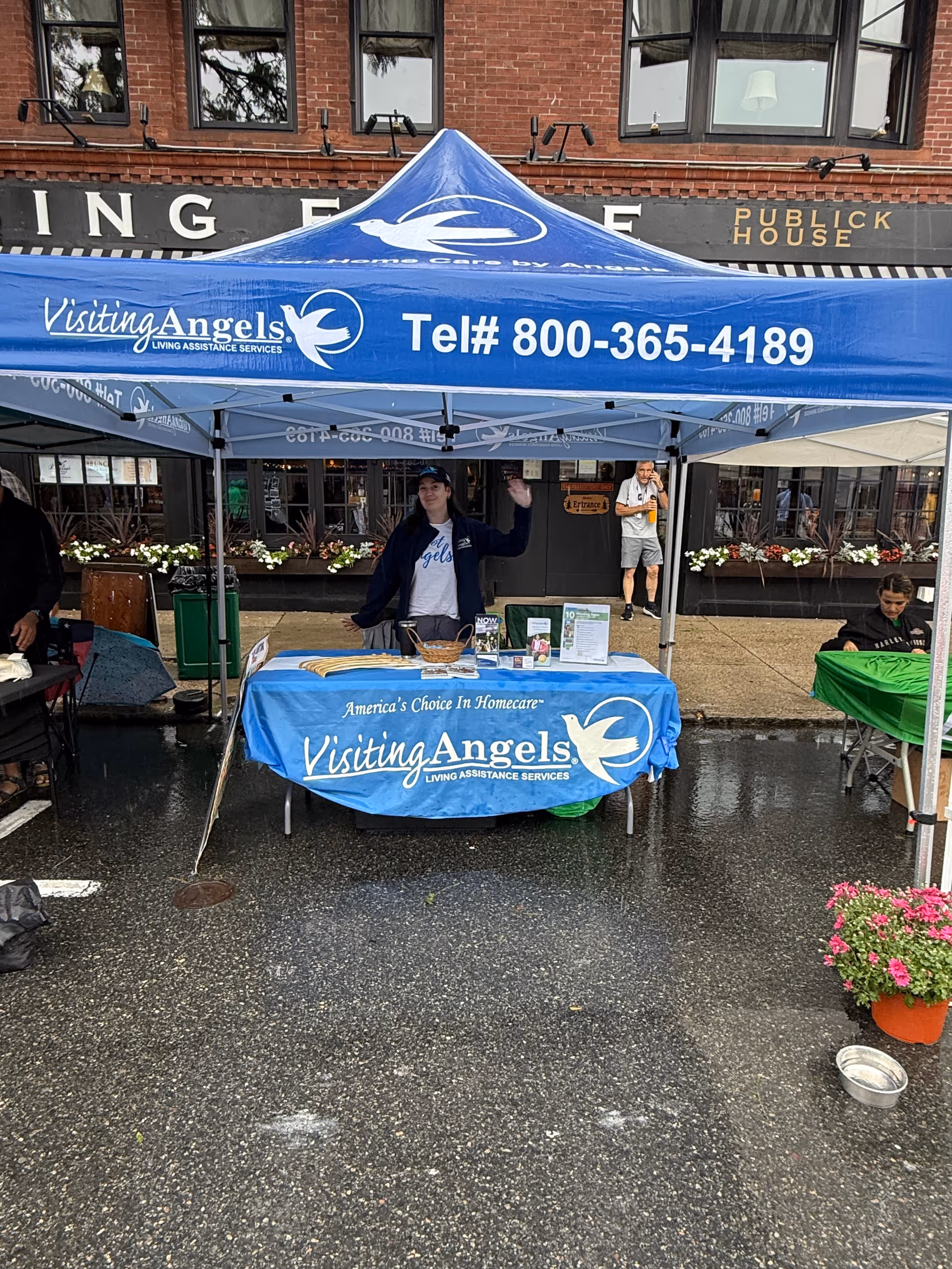 Blue Visiting Angels tent and table at an outdoor event in front of a brick building.