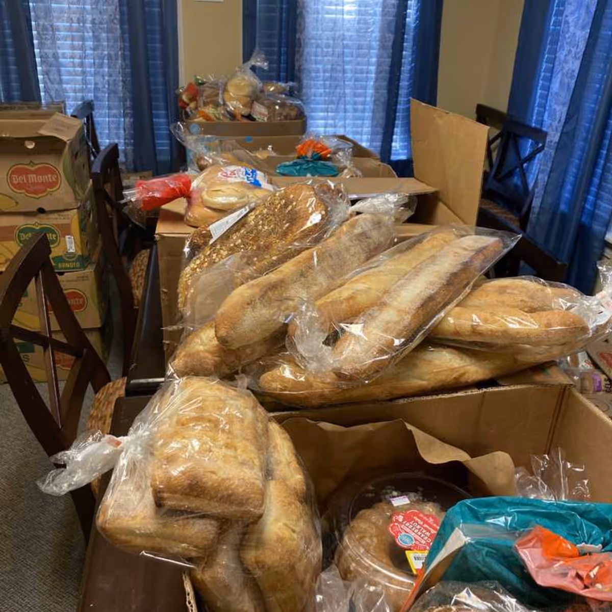 Bread loaves in plastic bags stacked on tables and boxes in a dining room.