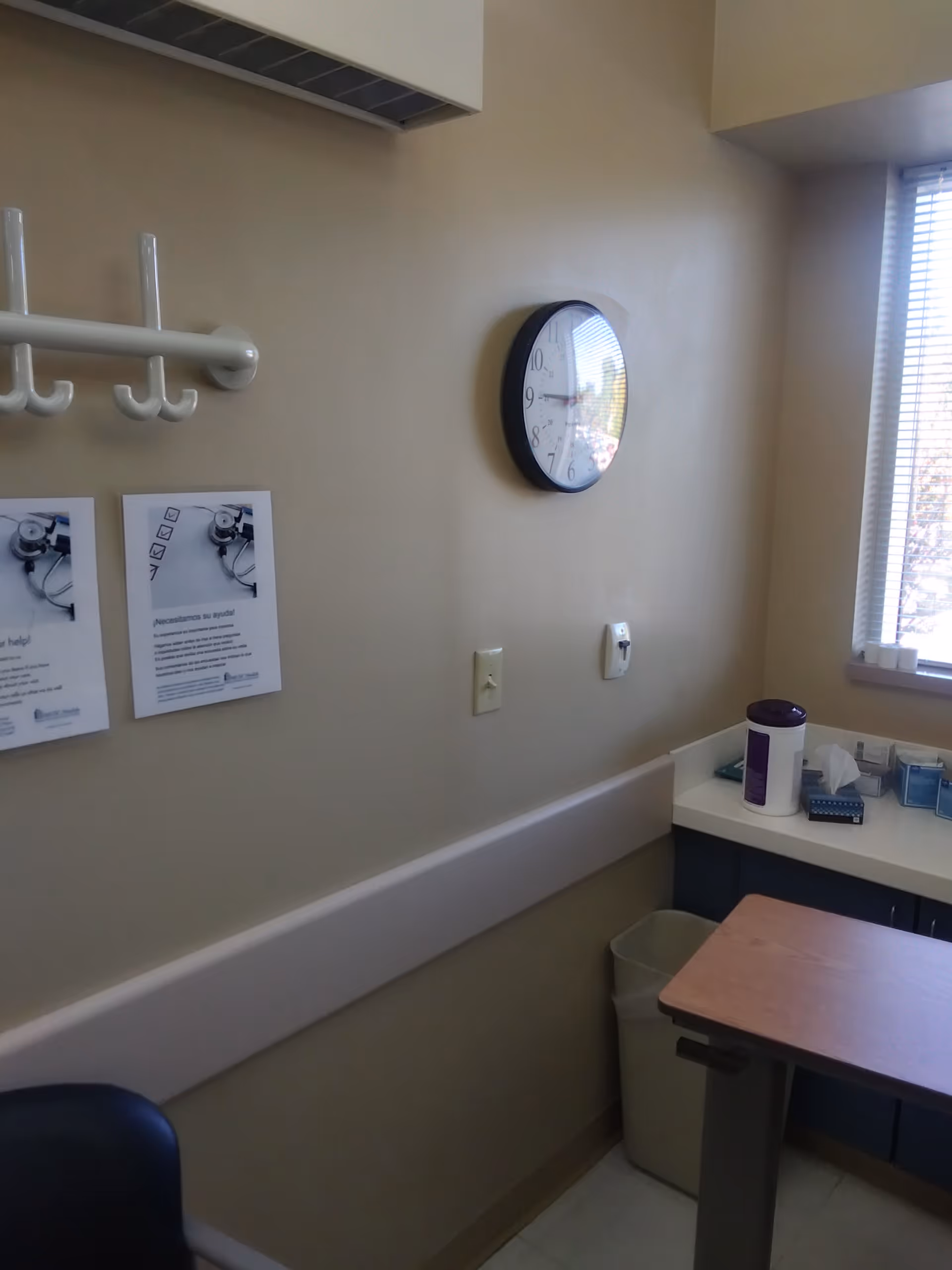 Corner of a medical examination room with clock, hooks, and a small desk near a window.