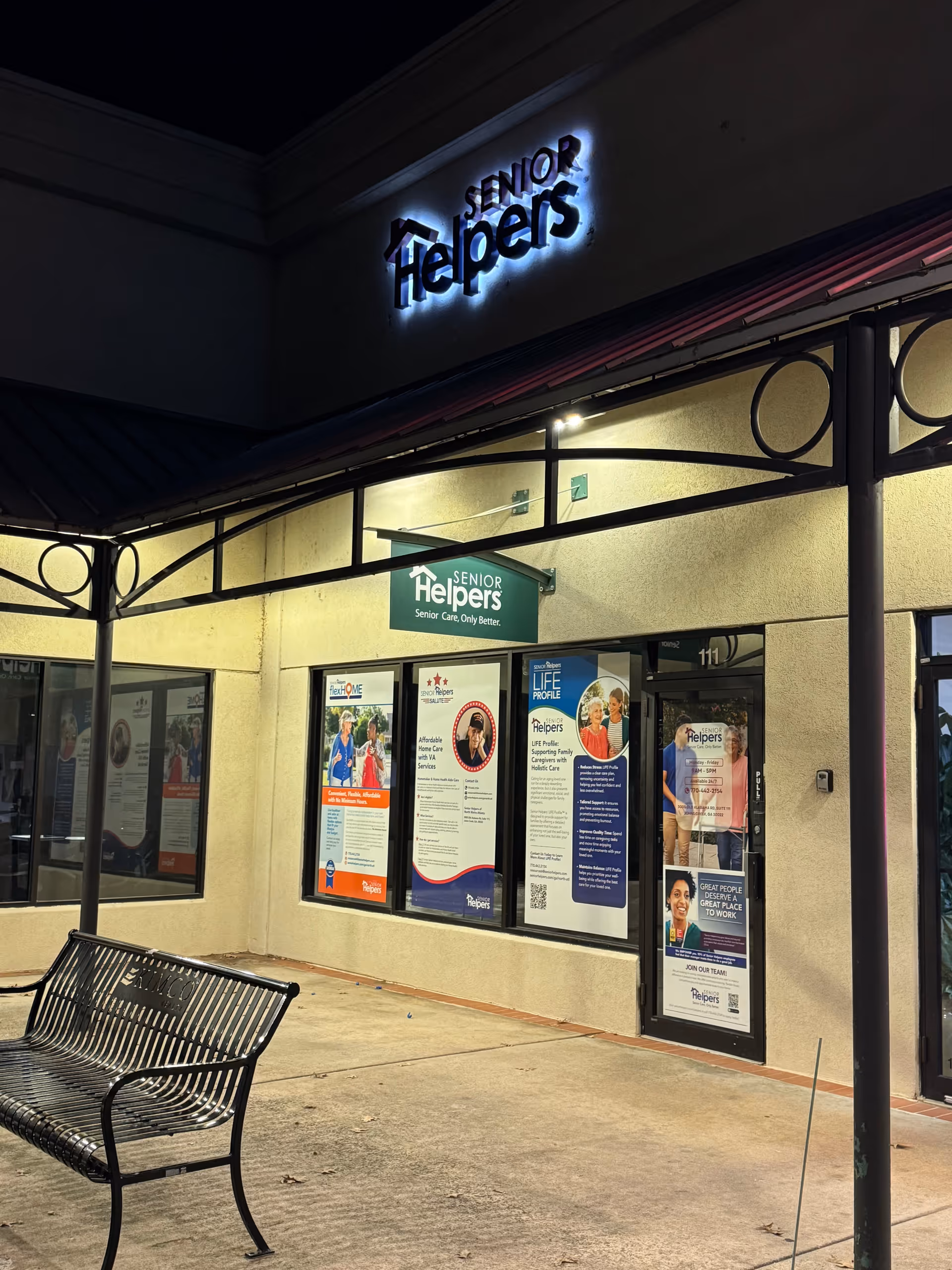 Exterior at night of Senior Helpers facility with illuminated sign, posters in window, and a bench.