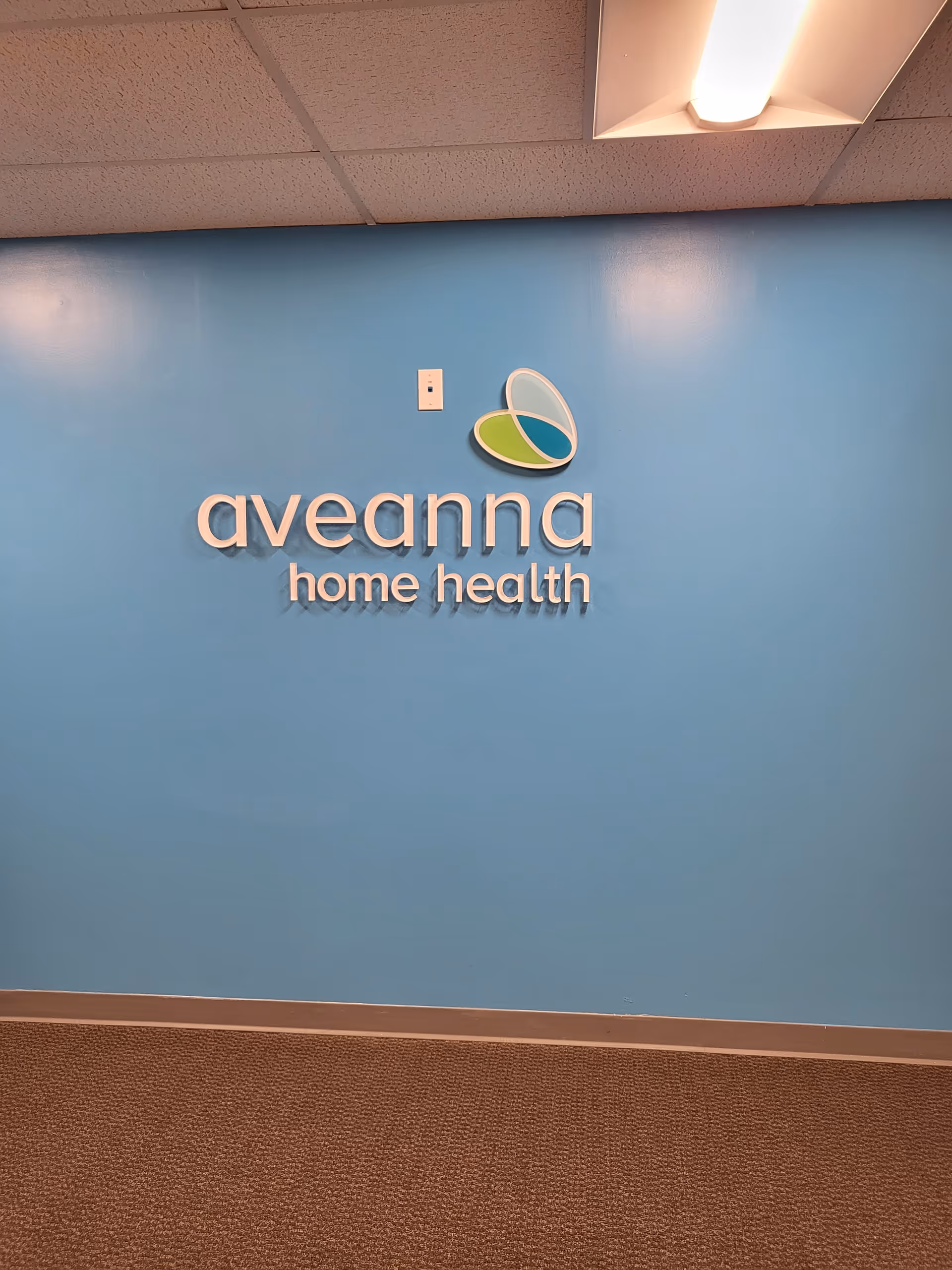 Blue interior wall displays Aveanna Home Health logo beneath a ceiling light.