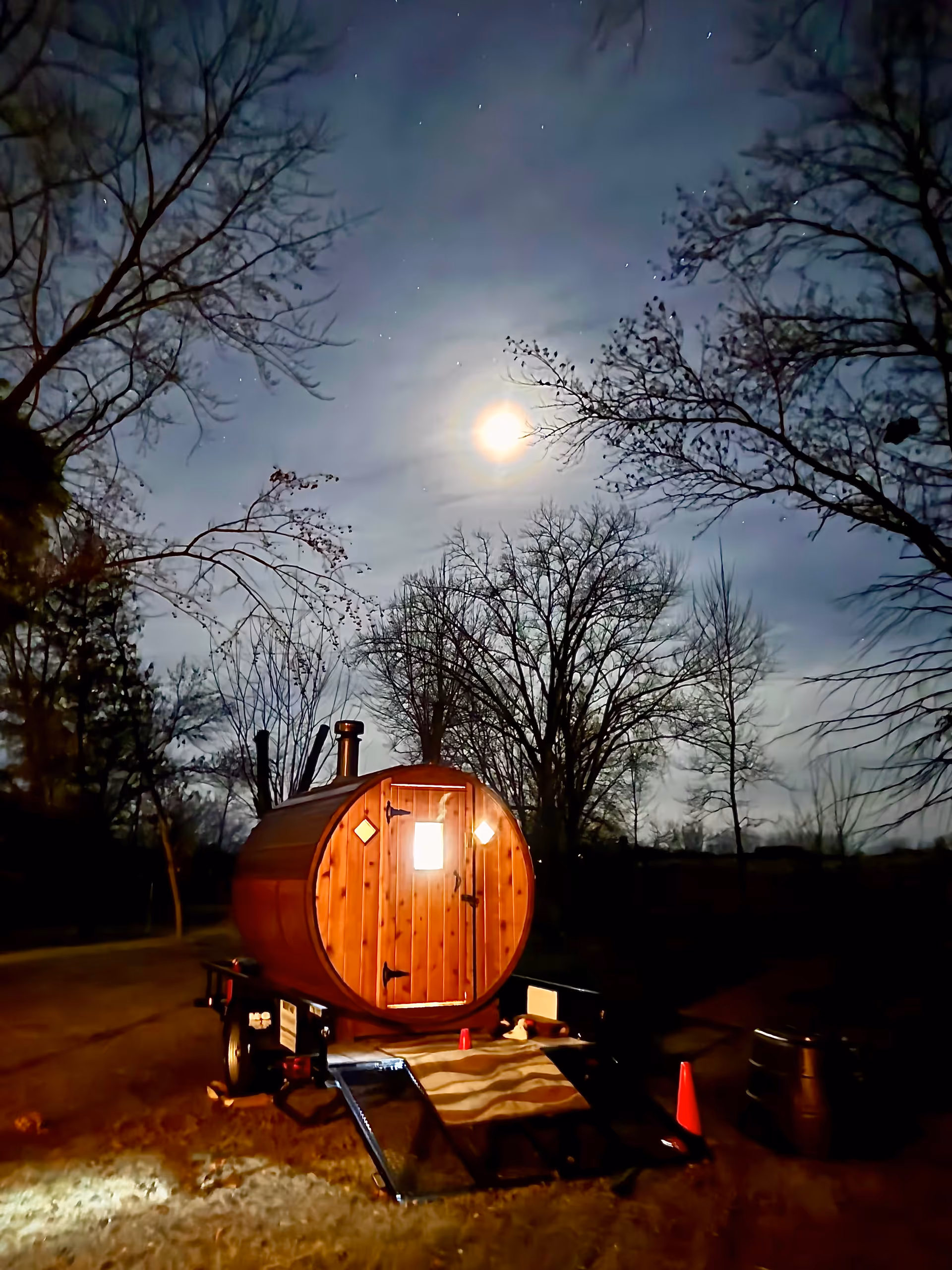 Nighttime outdoor wooden barrel sauna on trailer, surrounded by bare trees under moonlit sky.