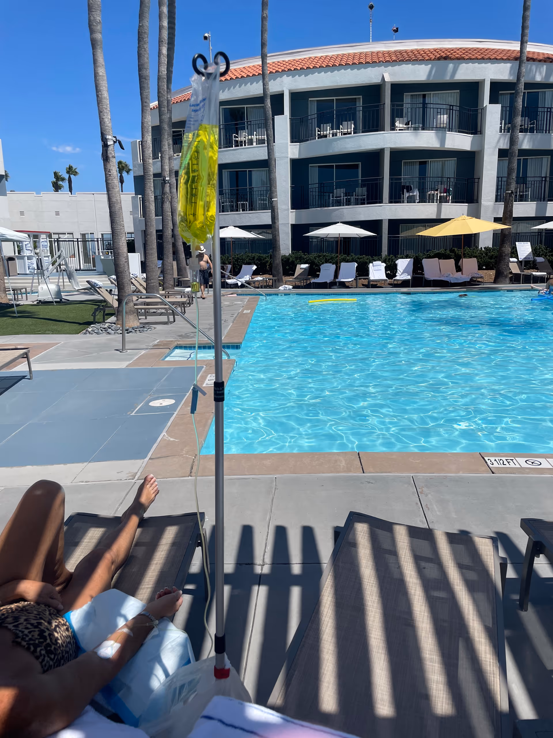 Outdoor pool area with lounge chairs, palm trees, and an IV stand beside a sunbathing resident.