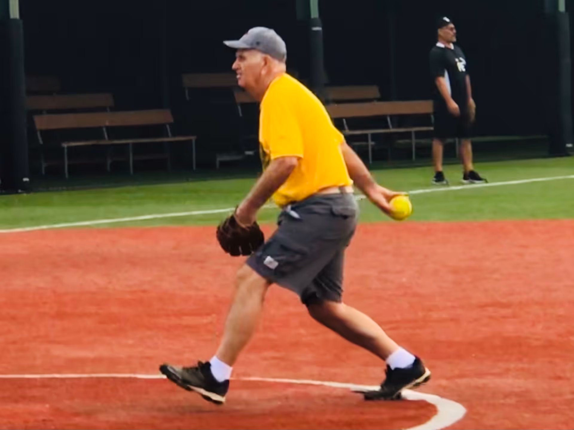 Older man in yellow shirt runs with glove, holding a ball on a red softball field.