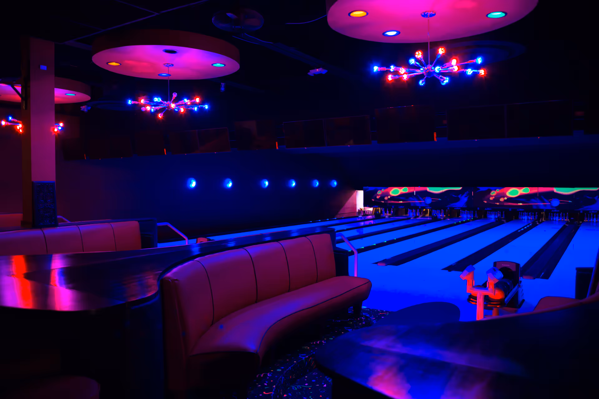 Dimly lit bowling alley with neon lights, multiple lanes, and curved seating in foreground.