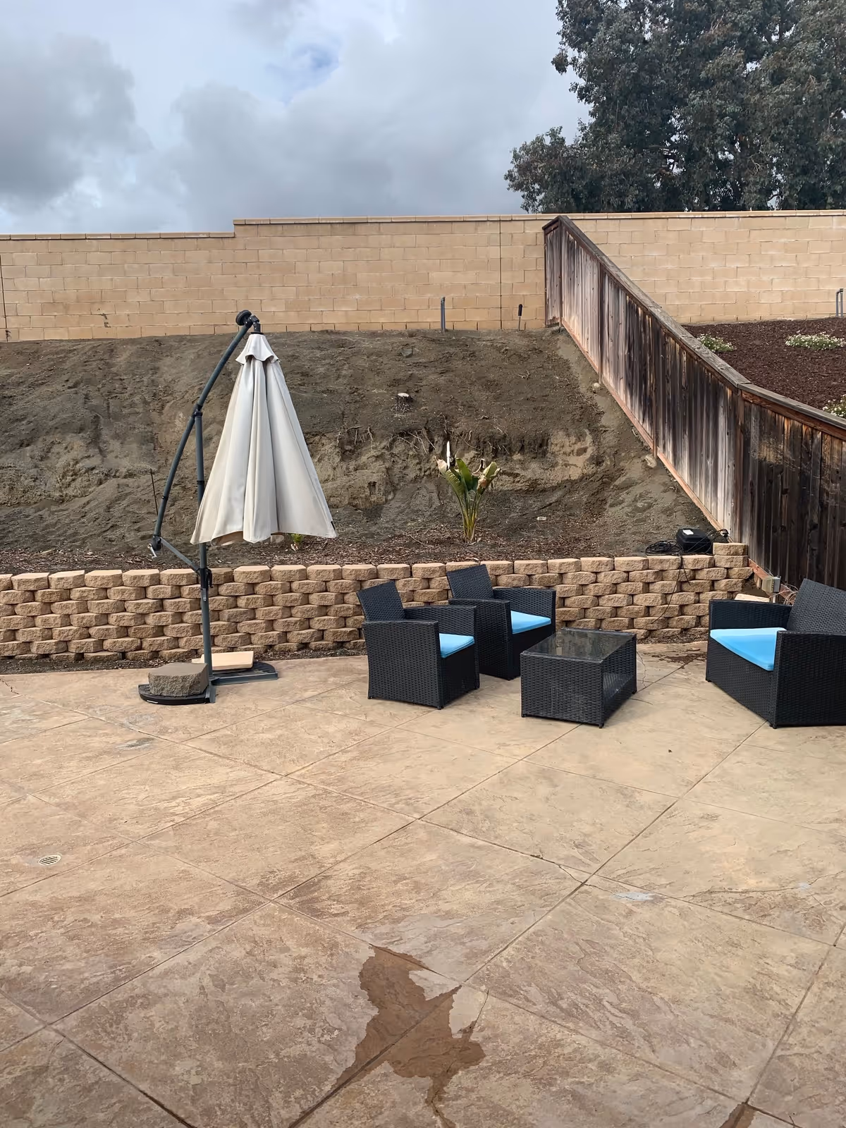 Outdoor patio with wicker seating, a glass table, and an overturned umbrella against a retaining wall.