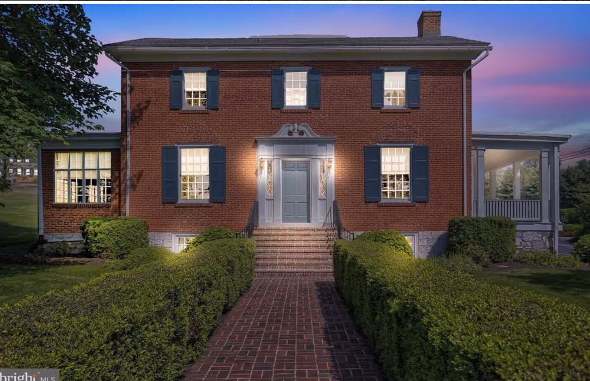 Red brick two-story building with blue shutters and a brick walkway lined by trimmed hedges.