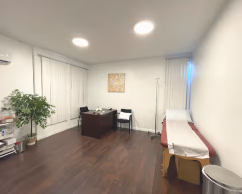 Empty clinic examination room with a desk, chairs, exam bed, and a potted plant.