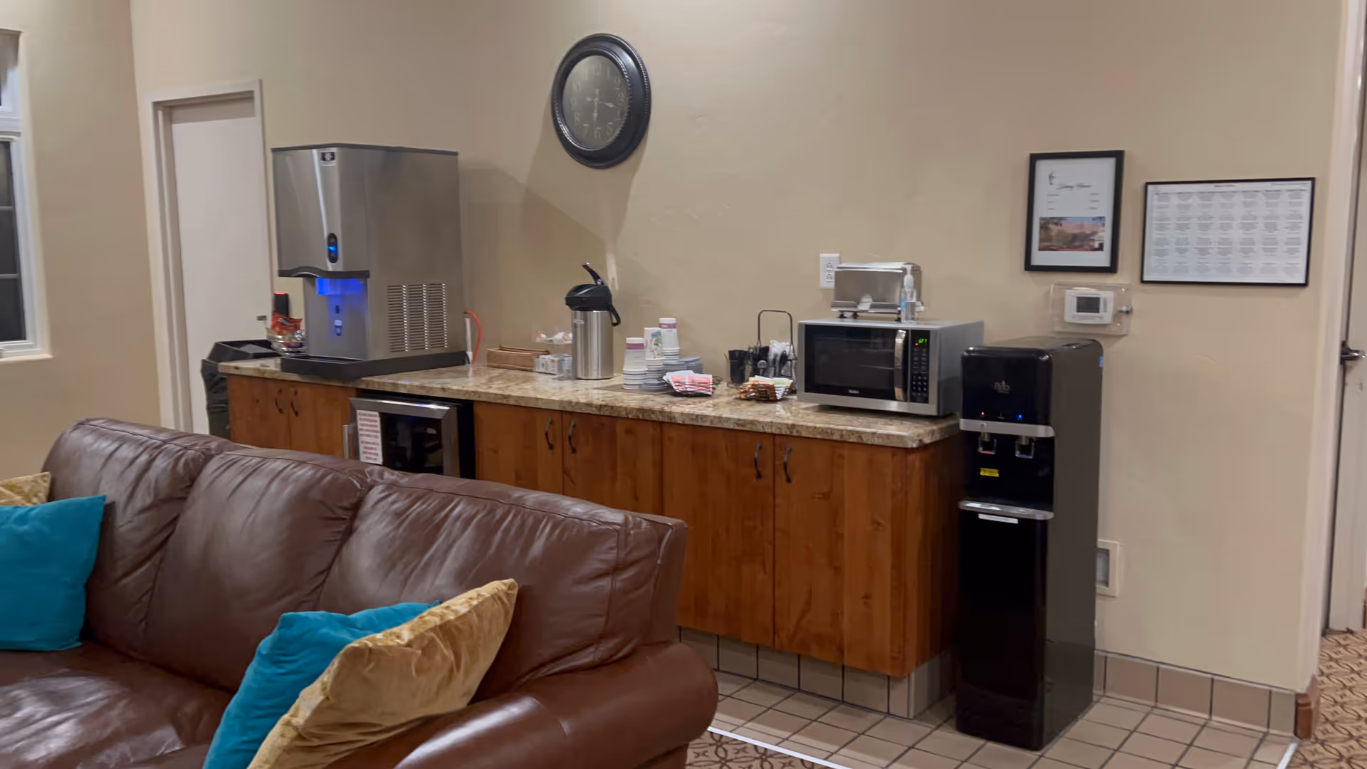 Brown leather sofa with teal pillows faces a kitchenette and coffee station in a common lounge.