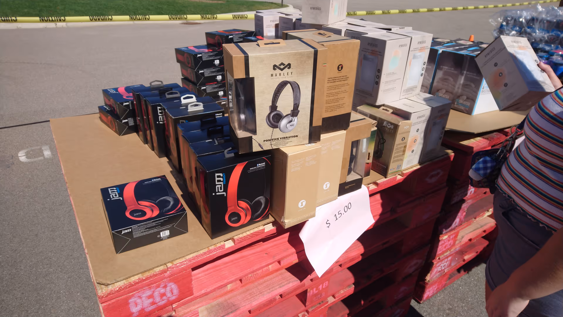 Pallets stacked with boxed electronics and headphones outdoors beside caution tape.
