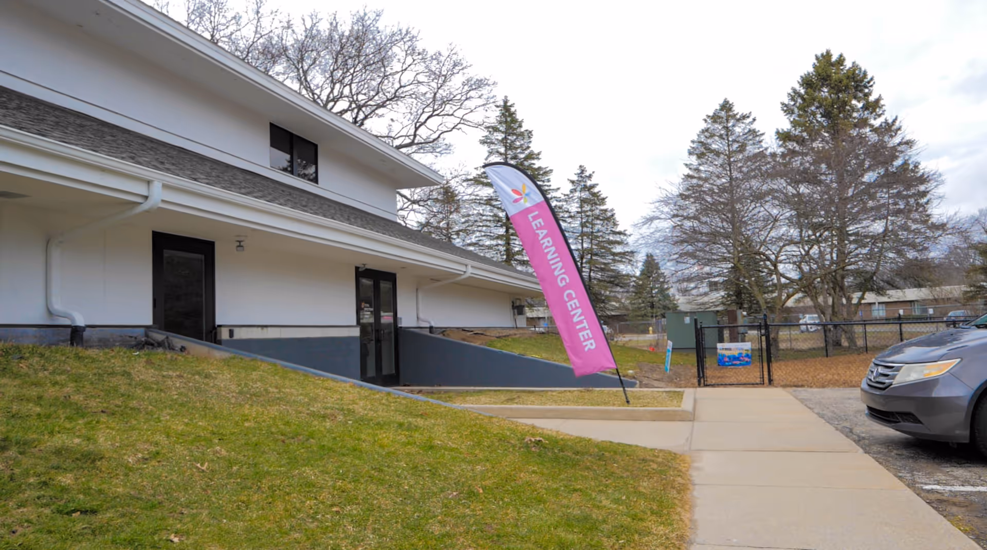 Exterior view of a white facility building with a ramp, sidewalk, fence, and pink banner.
