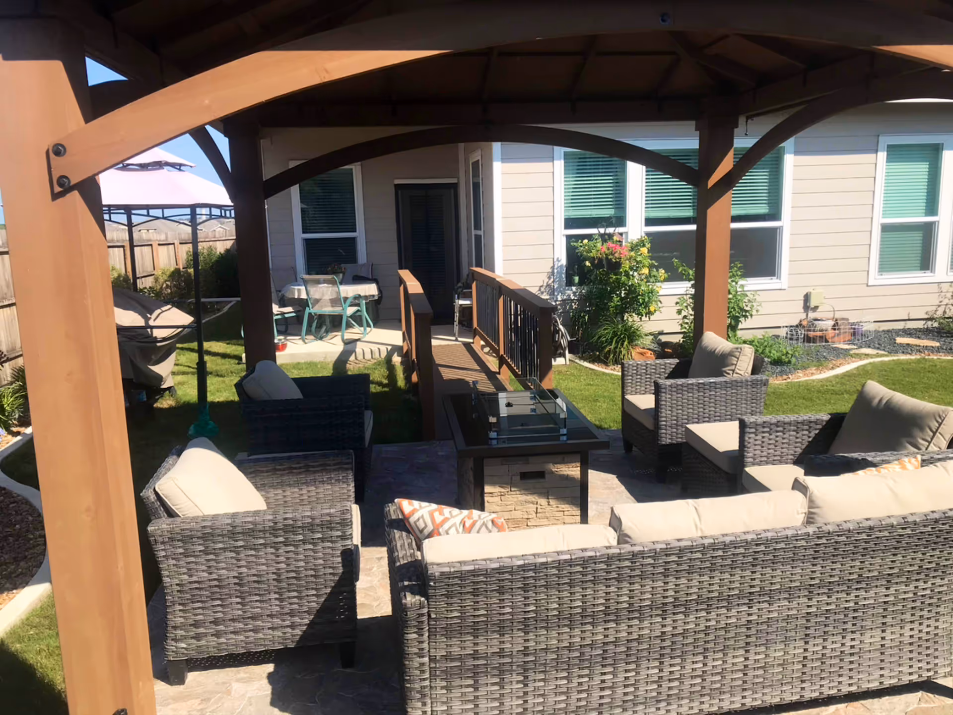 Outdoor patio area with woven furniture, cushions, and a pergola in a residential backyard.