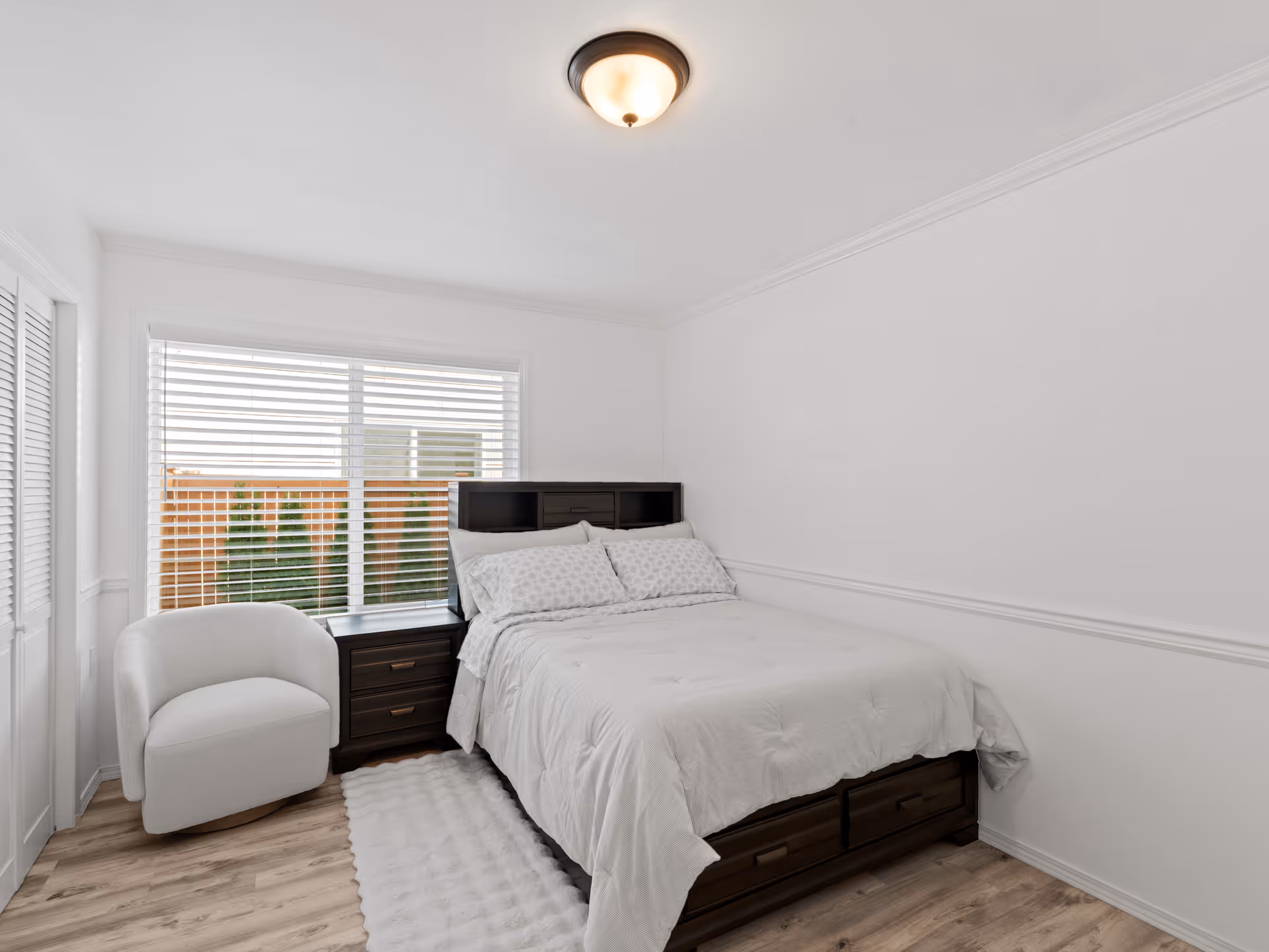 Bright bedroom with white bedding, dark wood dresser and nightstands beside a window.