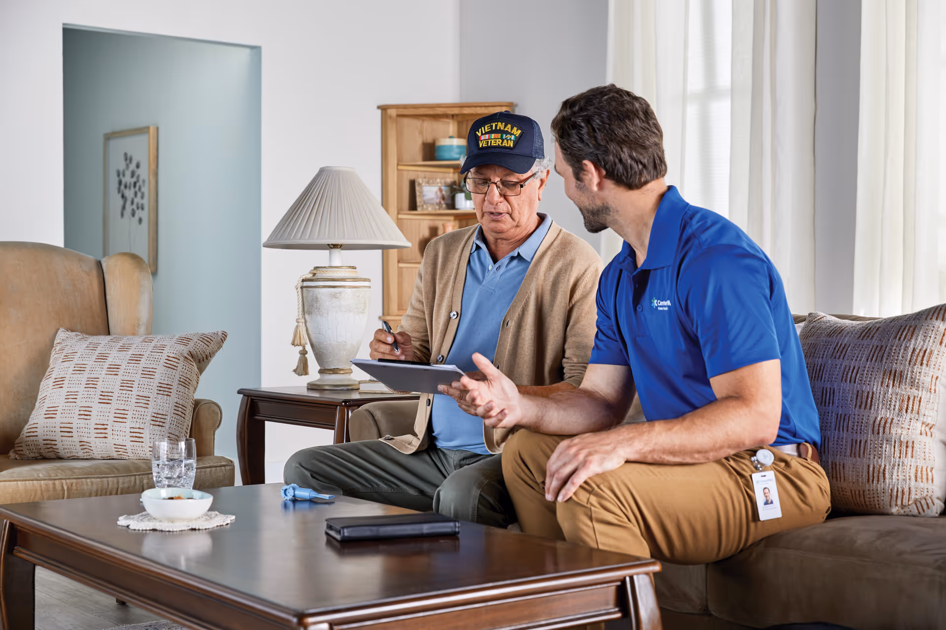 Two men sit on a couch in a living room, discussing a tablet.