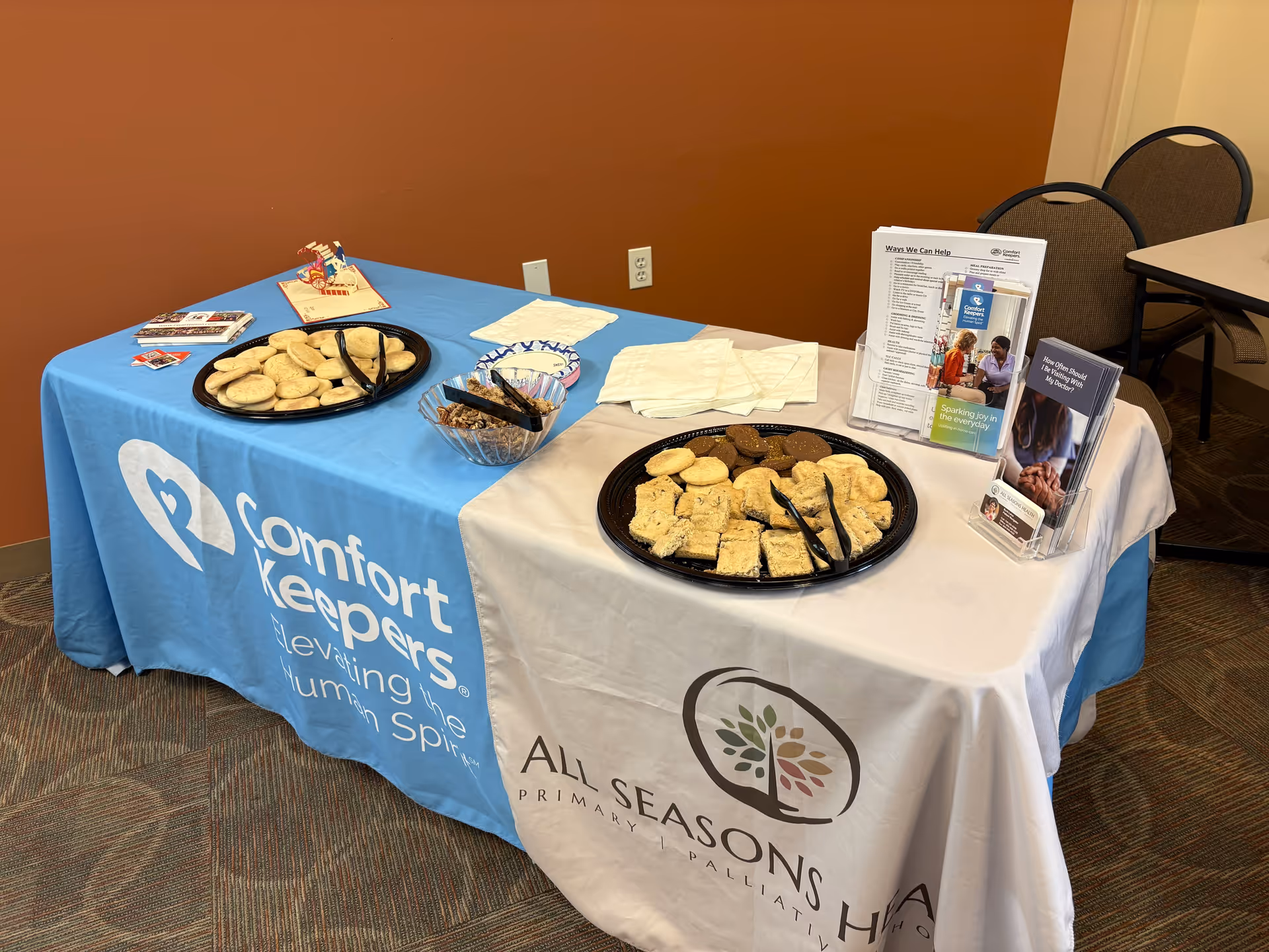 A table with cookies and brochures, covered with Comfort Keepers branding in a room.