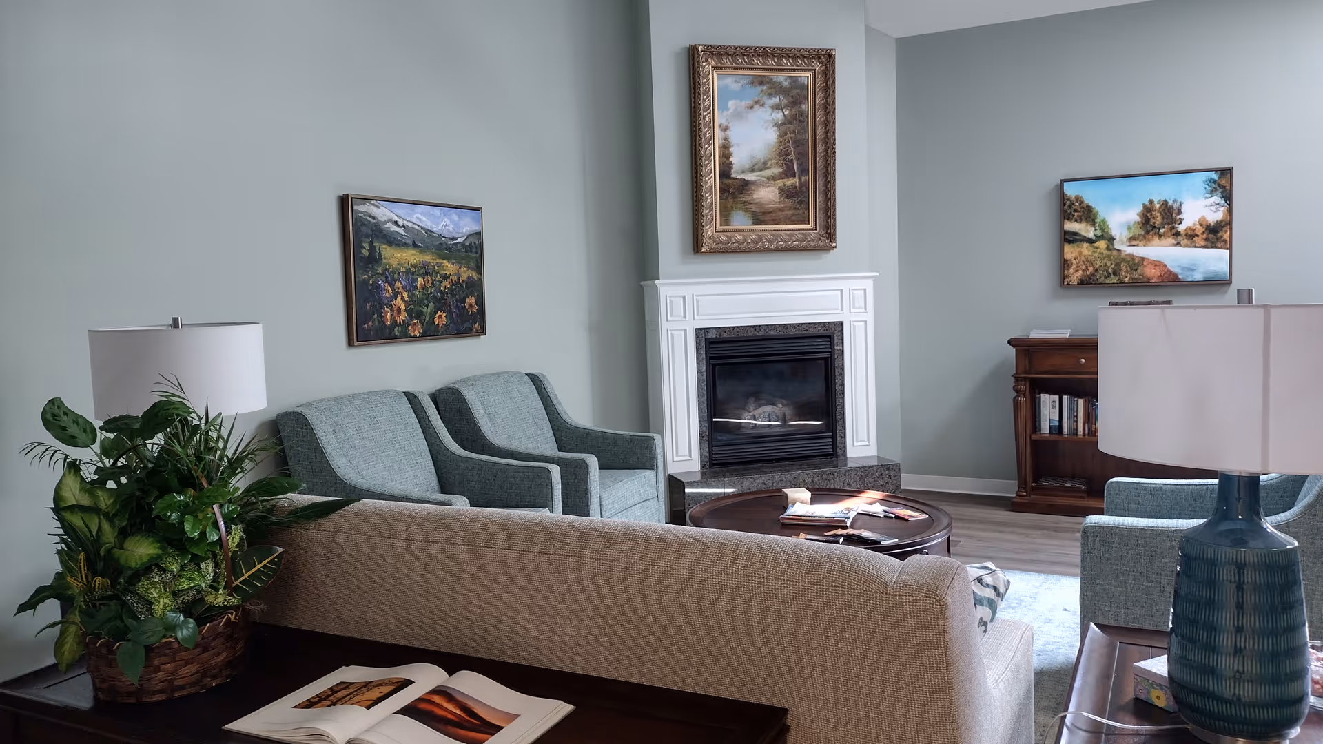 Light teal living room with fireplace, sofas, coffee table, plants, and landscape paintings.