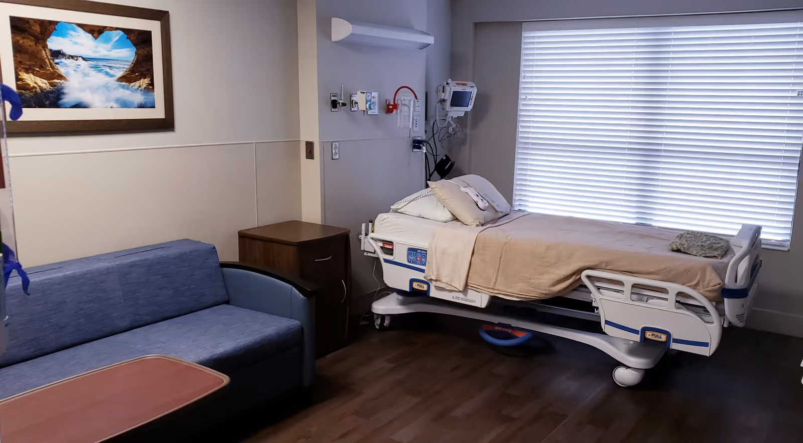 Hospital patient room with bed, medical equipment, a blue sofa, and blinds.