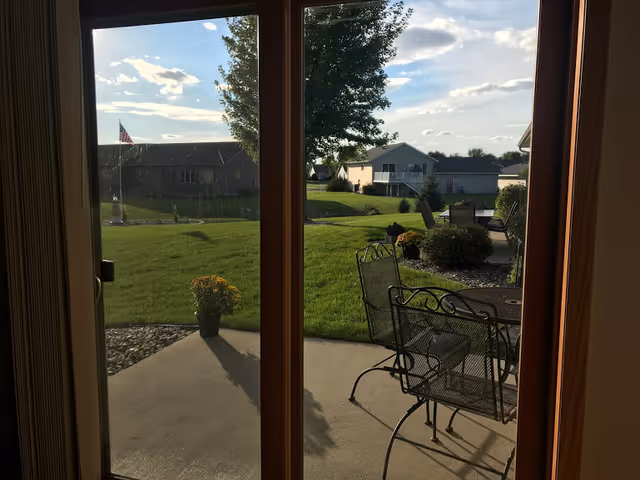 View through glass doors onto a patio with metal chairs and a green lawn.