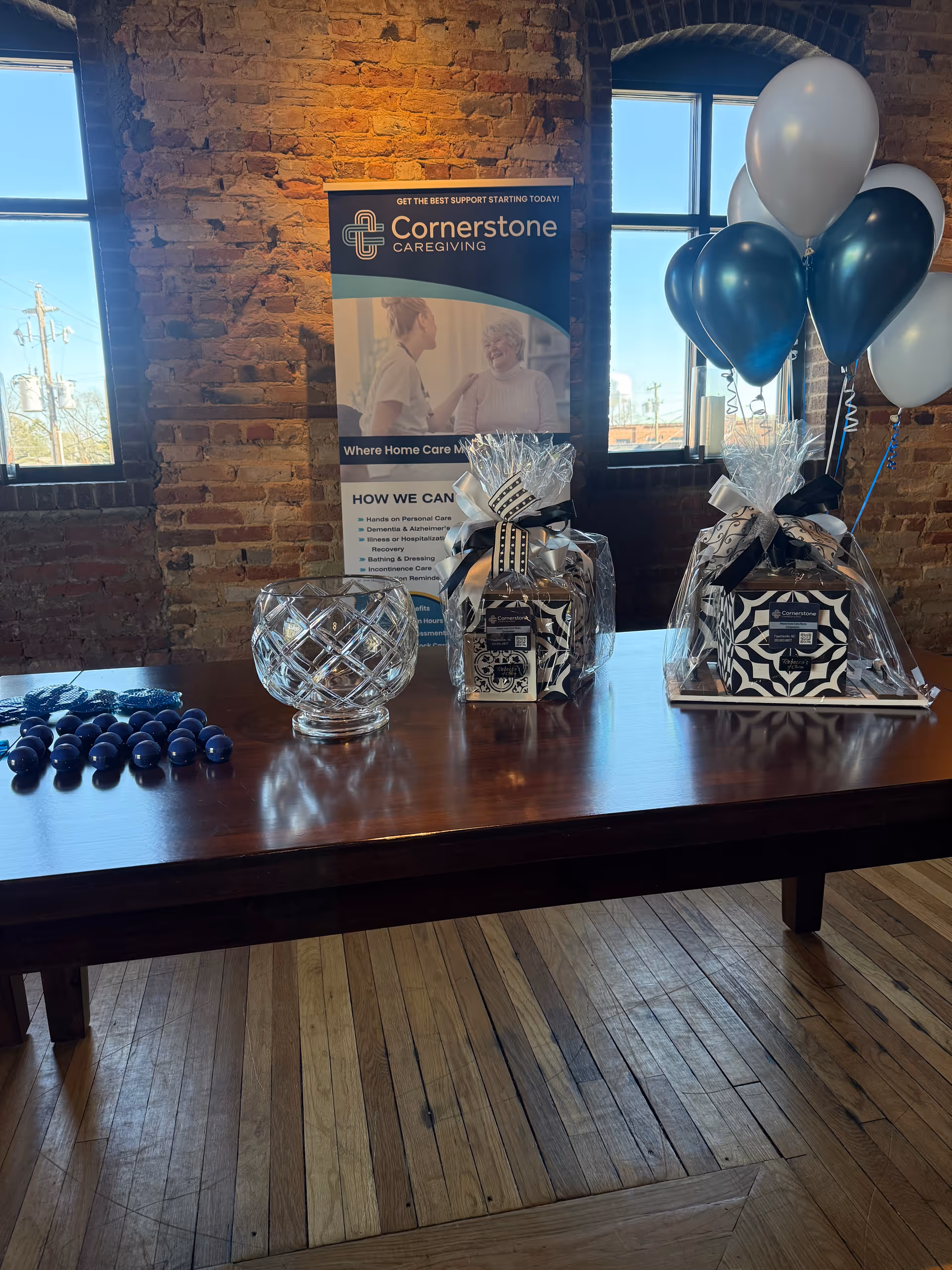 Indoor event setup with Cornerstone Caregiving banner, gift baskets, balloons, and a wooden table.