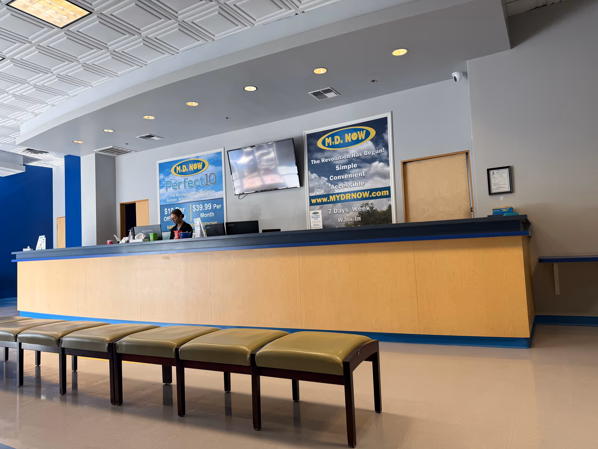 Lobby reception area with a long wooden counter, benches, and M.D. NOW posters.