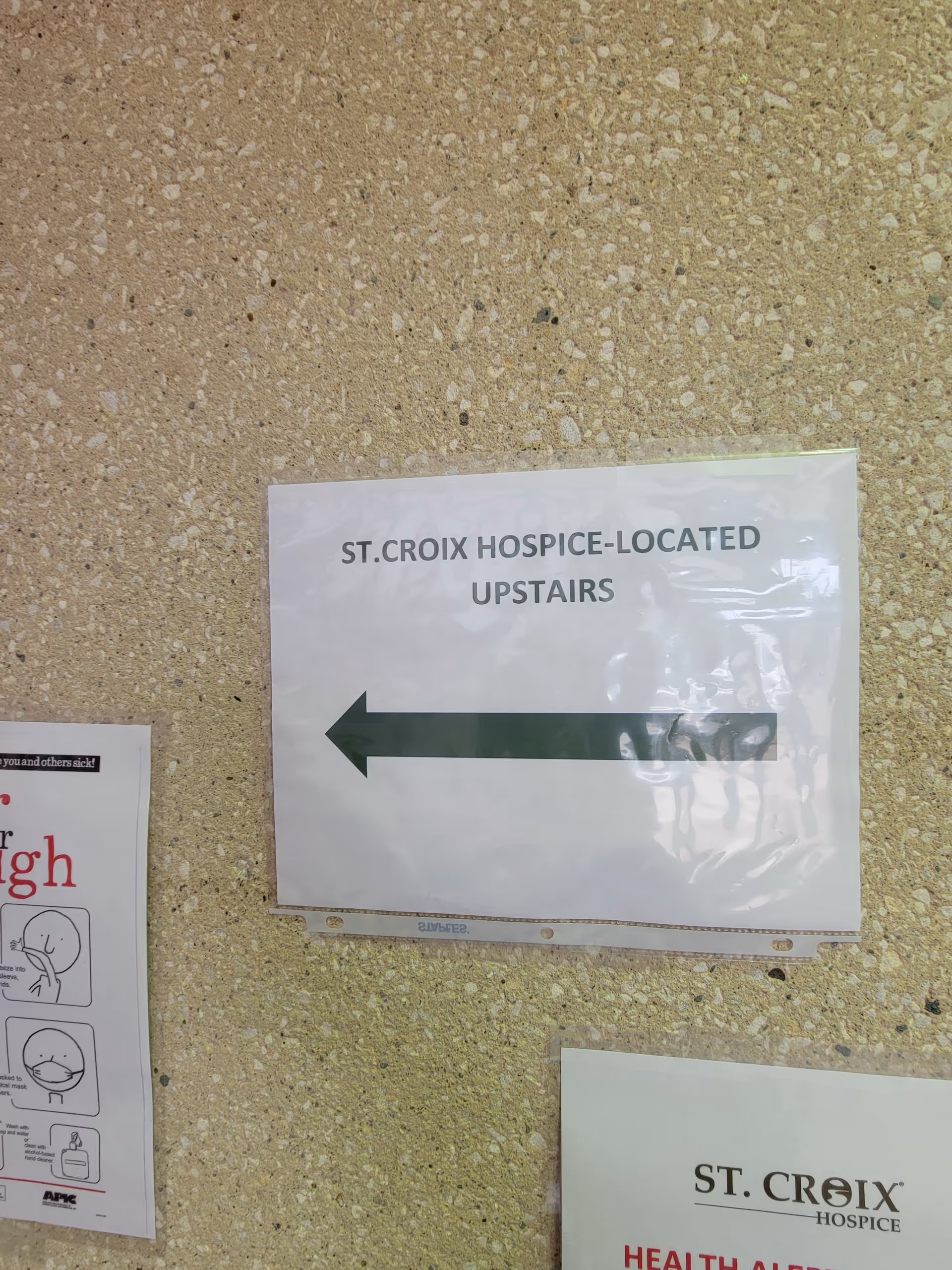 Sign on textured wall reading 'St. Croix Hospice Located Upstairs' with a left arrow.