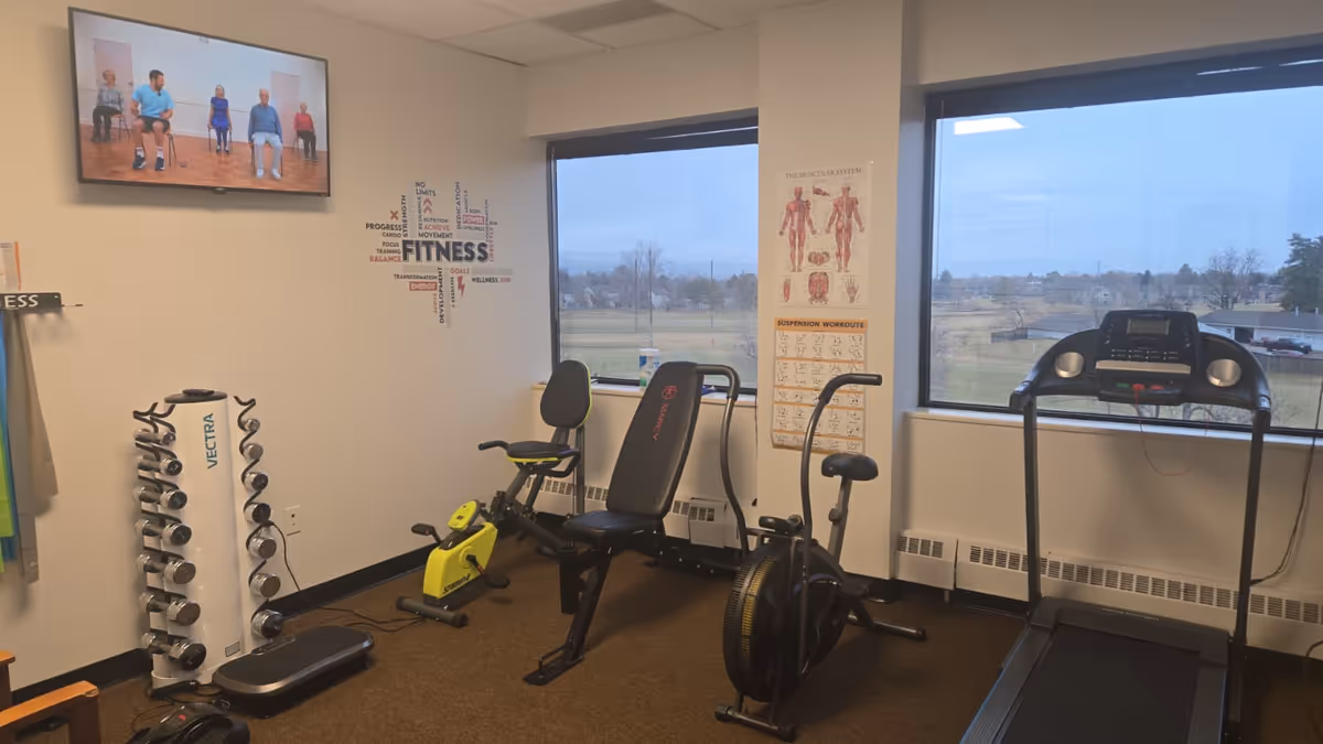 Senior living facility exercise room with treadmill, bike, weights, and posters by windows.