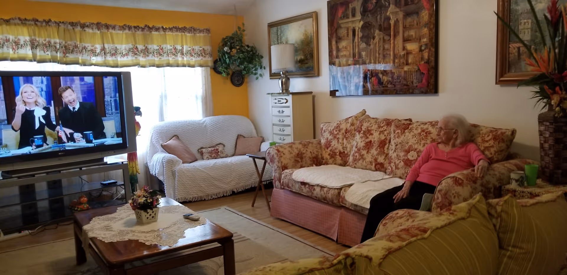 Senior woman relaxes on floral sofa in Glen Creek Villa living room with TV.