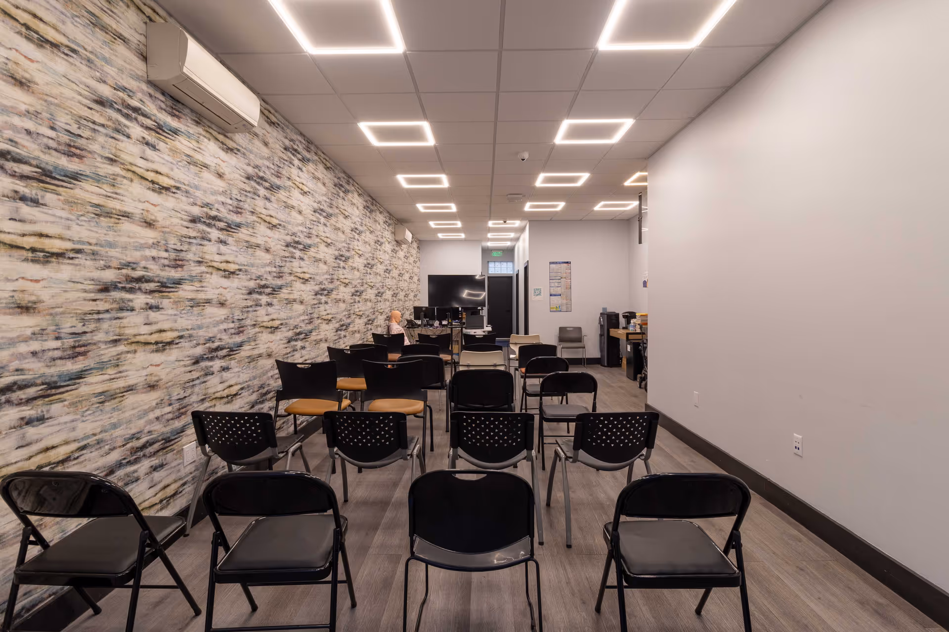 Long, well-lit conference room with rows of chairs facing a back workstation and patterned wall.