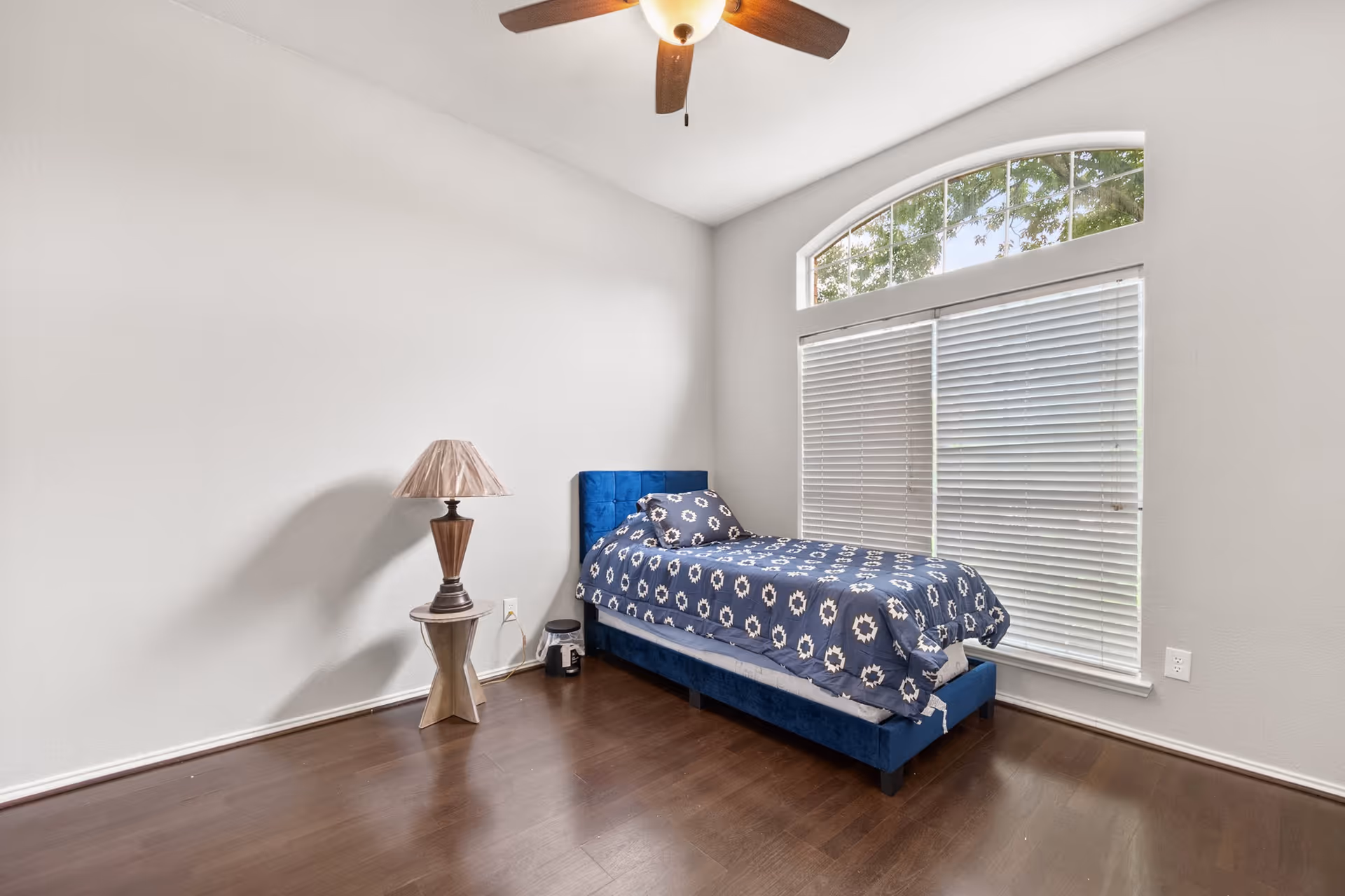 A clean bedroom with a blue upholstered bed, nightstand, lamp, and large window blinds.