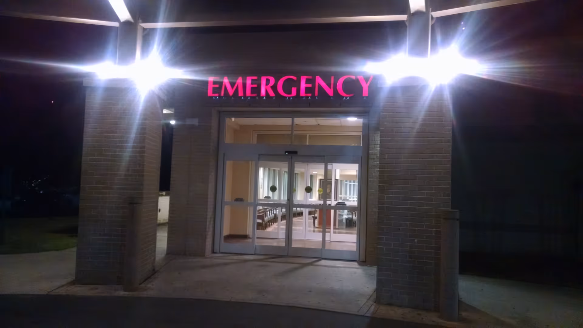 Nighttime exterior view of a hospital emergency entrance with a bright red EMERGENCY sign.