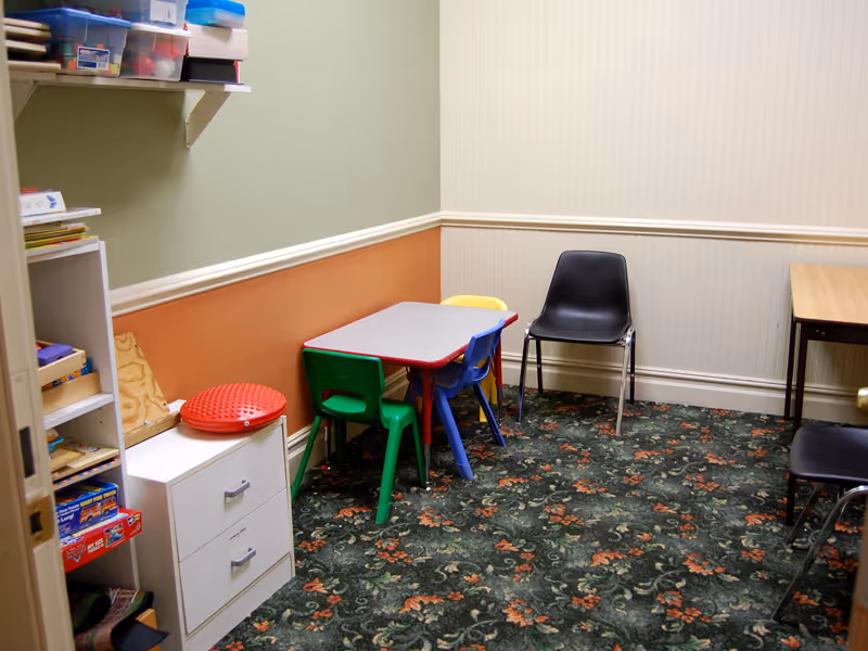 A small colorful children's table with chairs sits in a playroom beside shelves and patterned carpet.