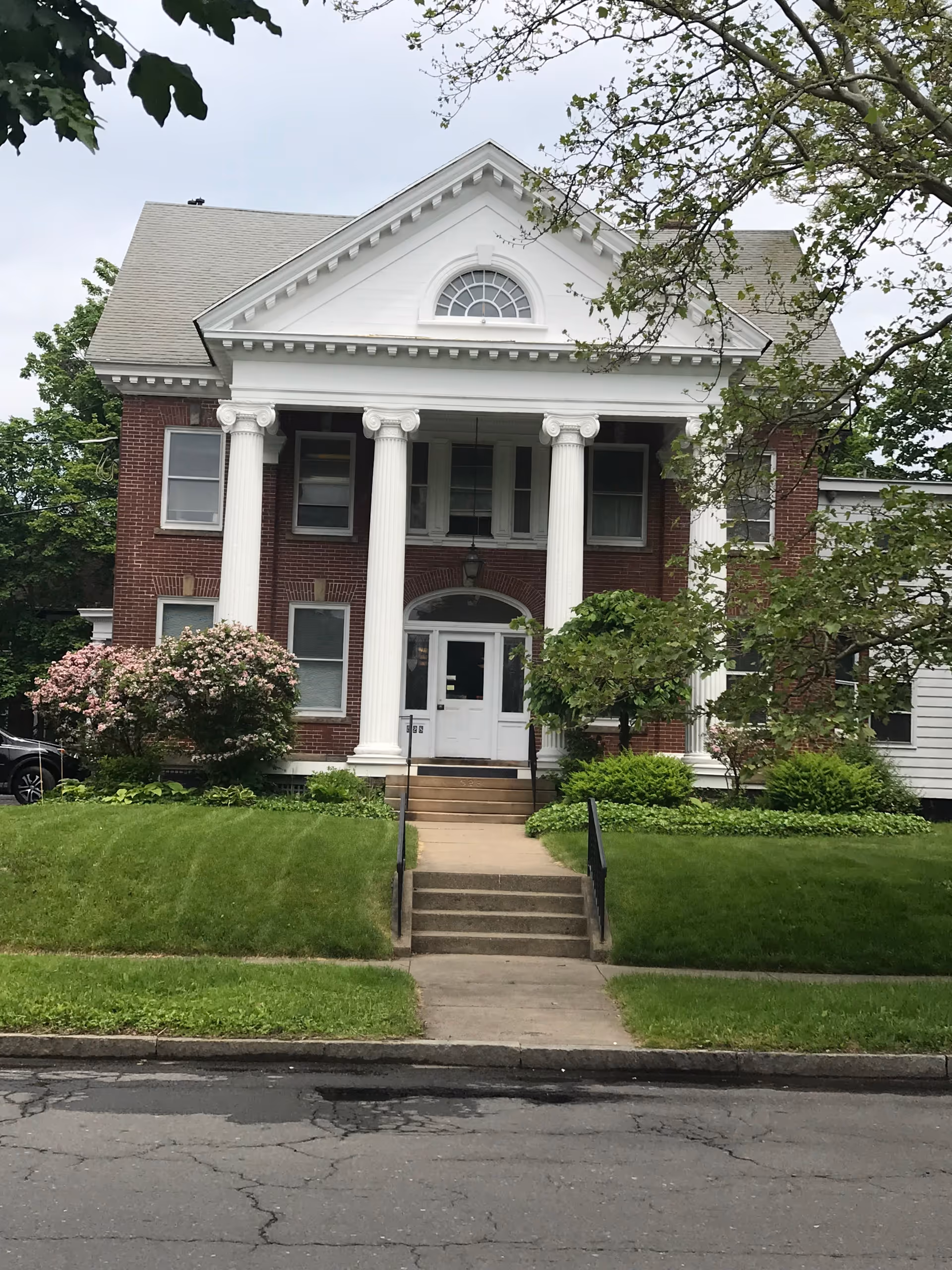 Front view of a brick building with white columns, steps, and green landscaping.