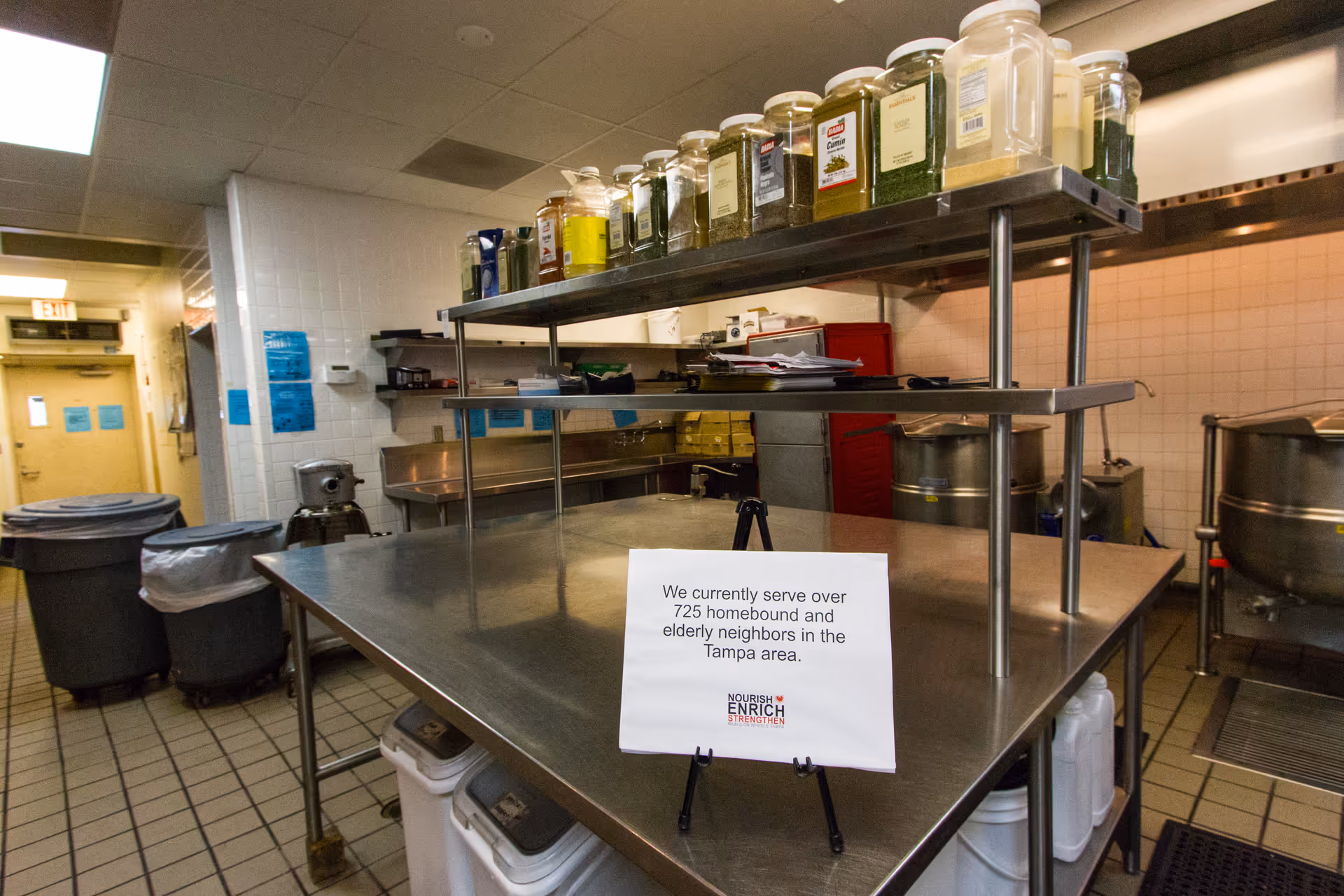 Industrial kitchen with stainless steel prep table, shelves of jars, and cooking equipment.