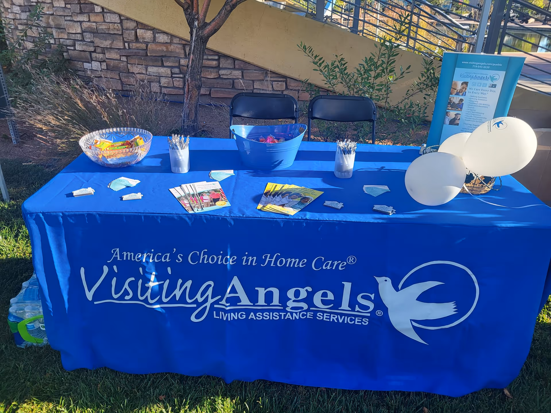 Outdoor table with blue Visiting Angels tablecloth, brochures, balloons, and snacks at a community event.