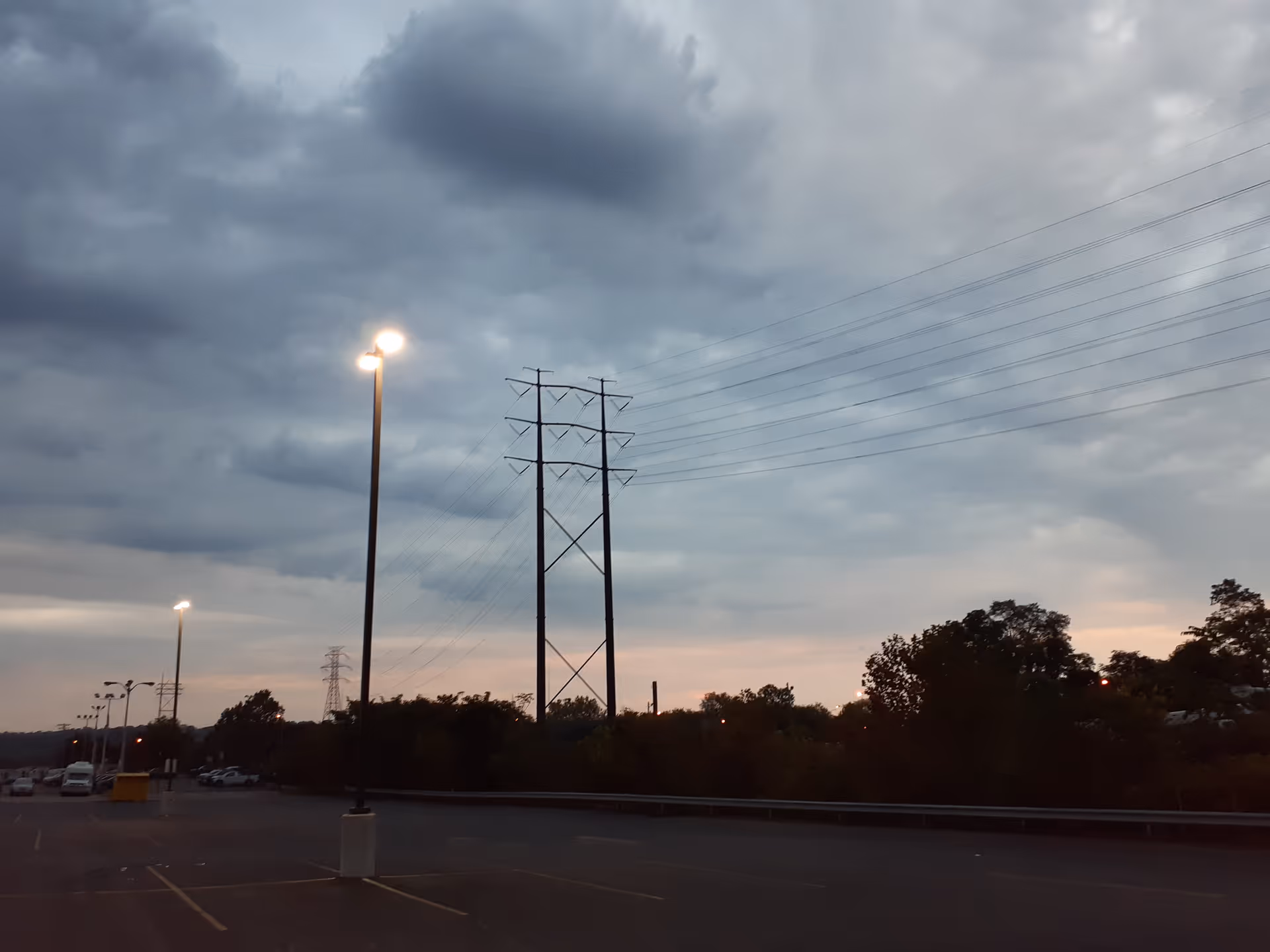 Empty parking lot at dusk with tall streetlights, power lines, and cloudy sky.