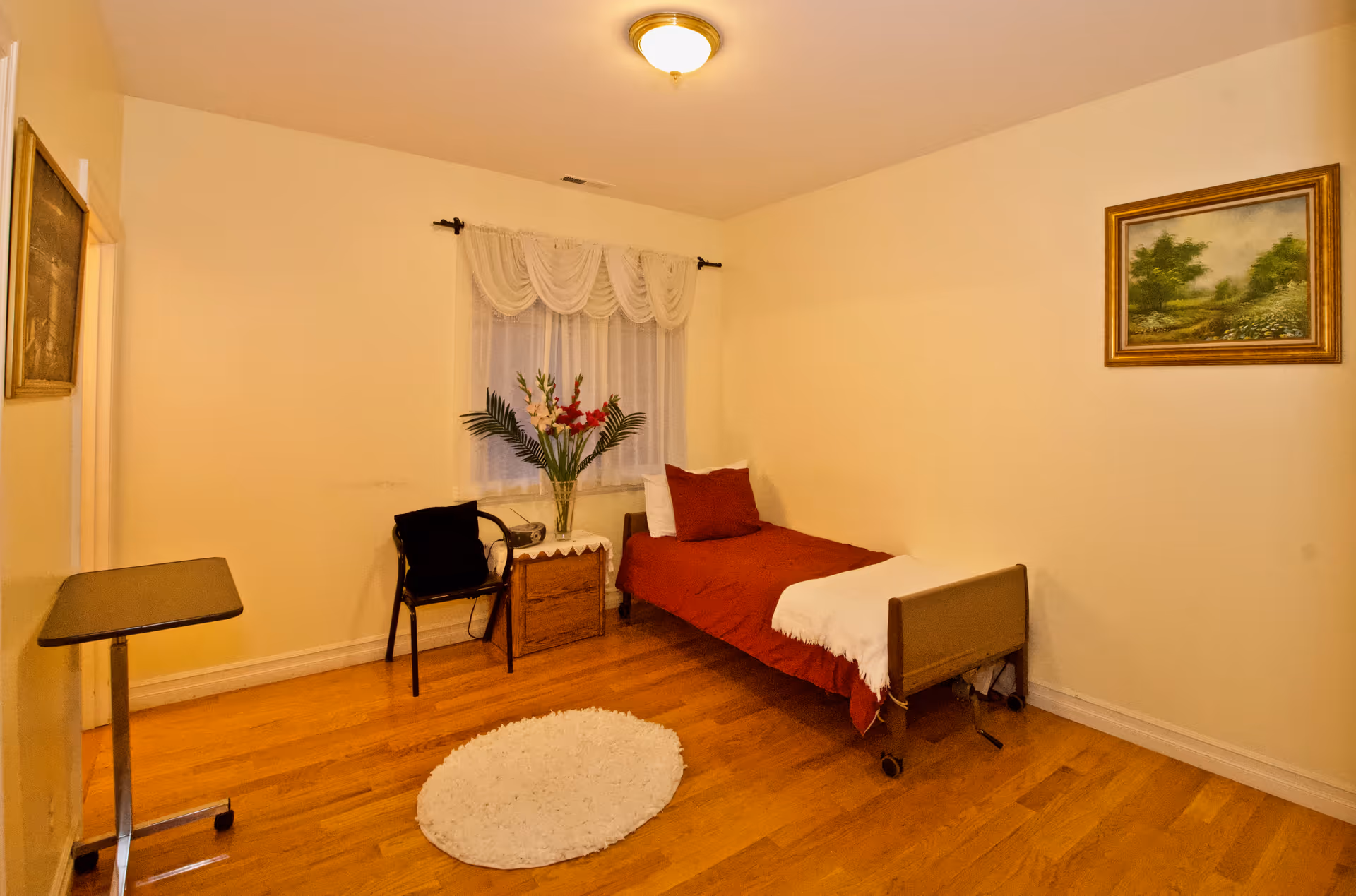 A simple senior bedroom with a single bed, nightstand, chair, vase of flowers, and rug.