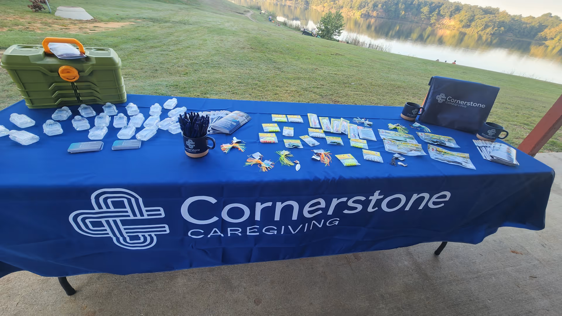 Outdoor table with Cornerstone Caregiving materials and freebies beside a grassy lakeside area.